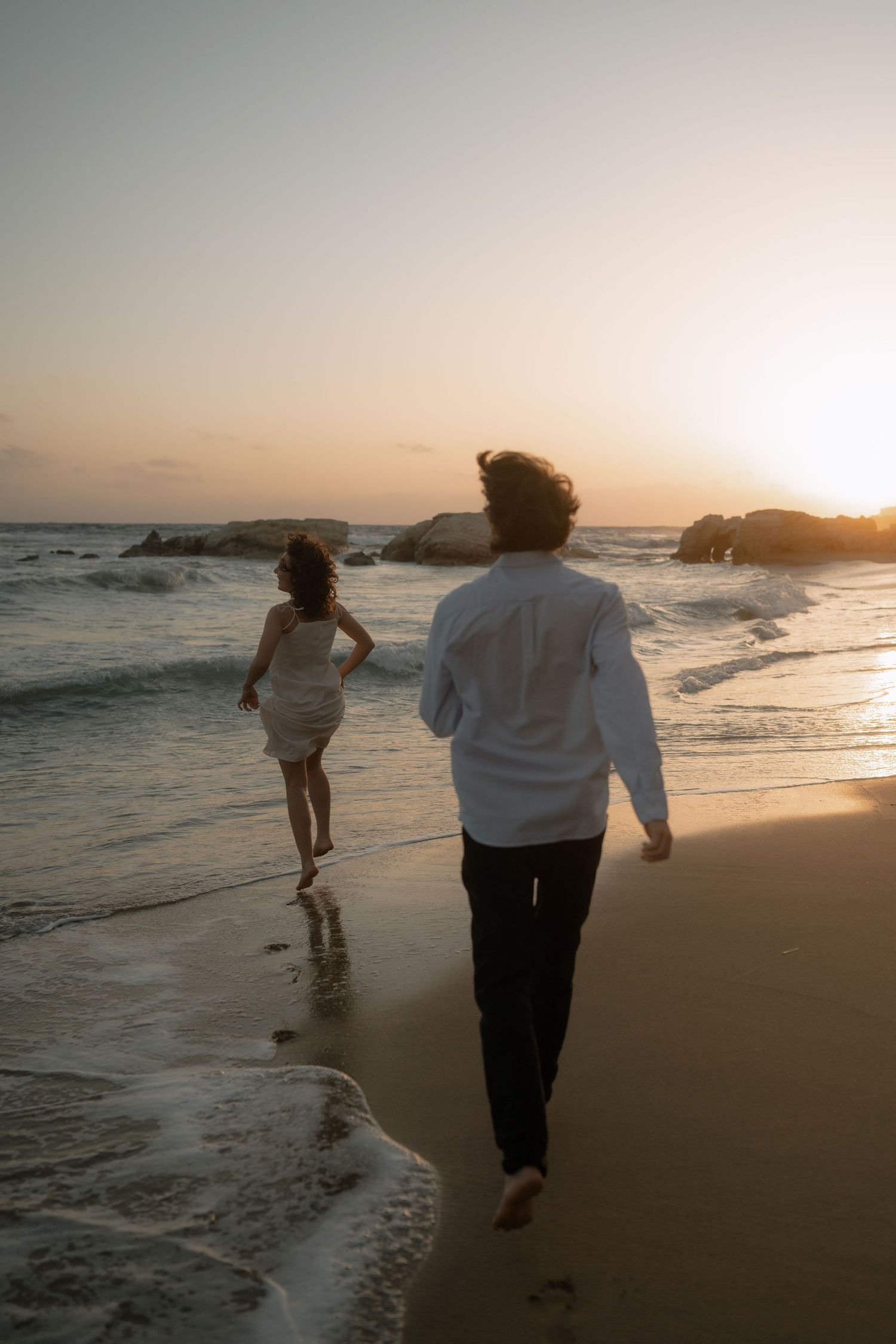 Couple in white outfits running barefoot on a sandy beach at sunset with ocean waves and rocks in background.