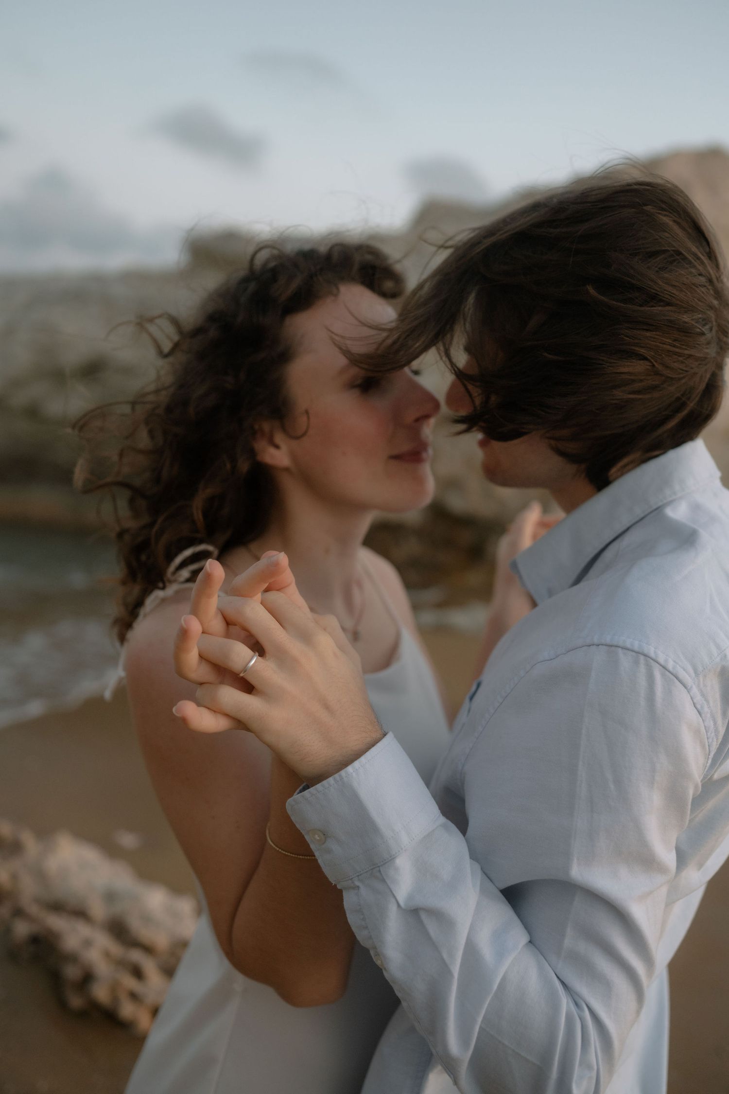 Romantic couple sharing an intimate moment on a beach, holding hands with waves and rocks in the background.