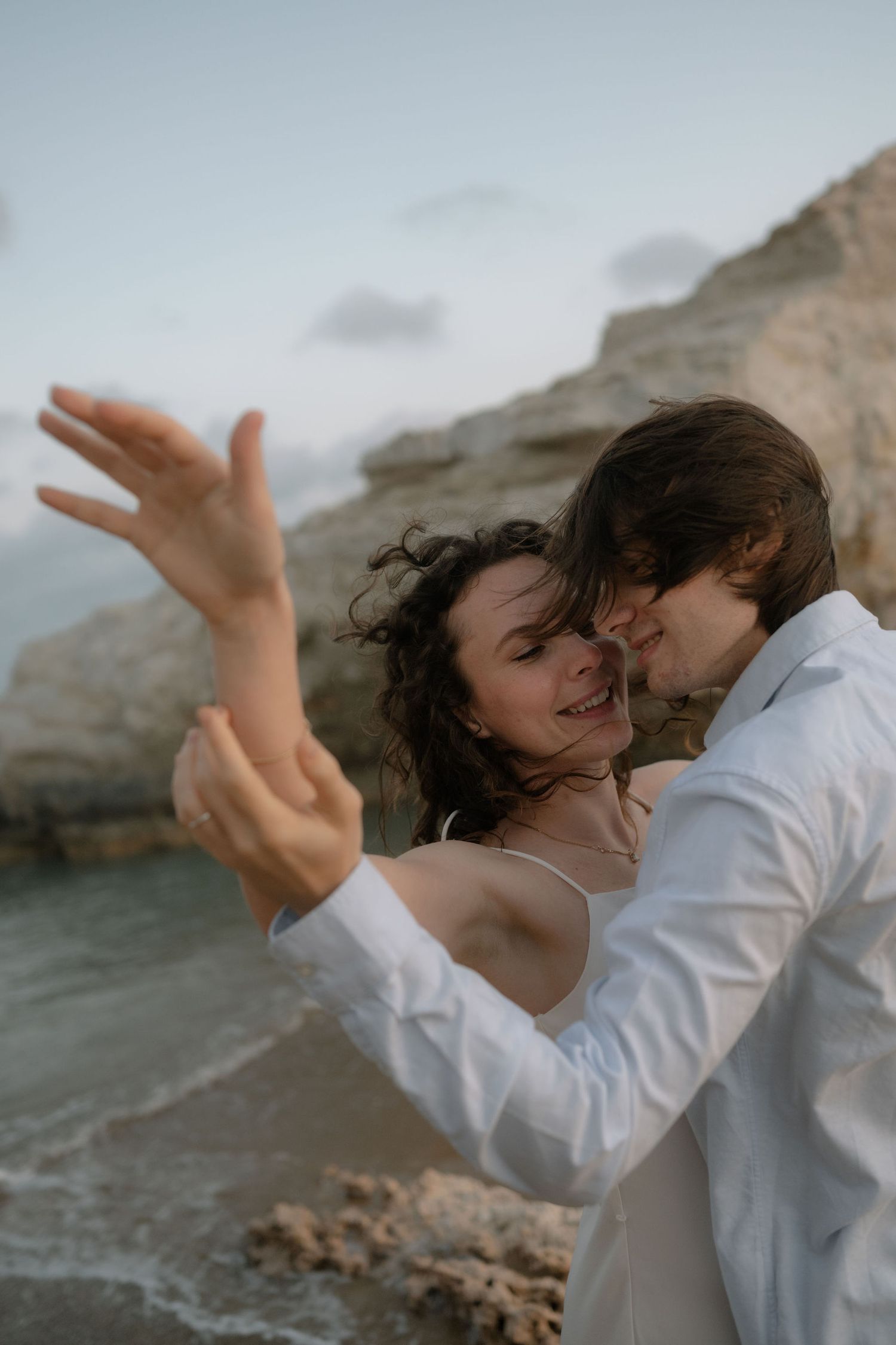 Bride and groom sharing tender moment on beach, woman smiling with raised hand, rocky cliffs in background.