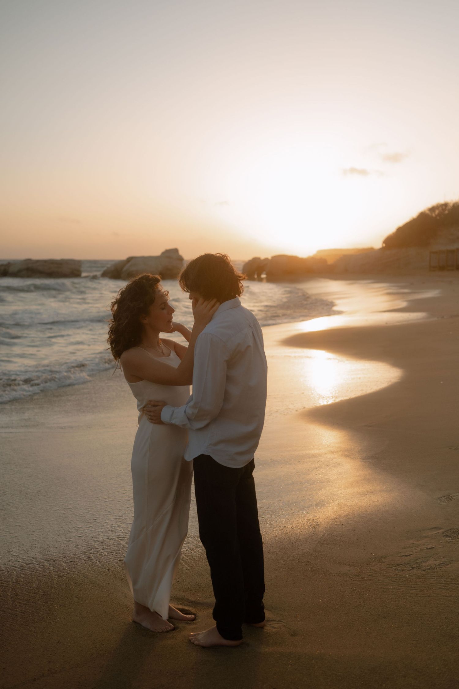 Couple embracing on a sandy beach at golden sunset with ocean waves in the background.