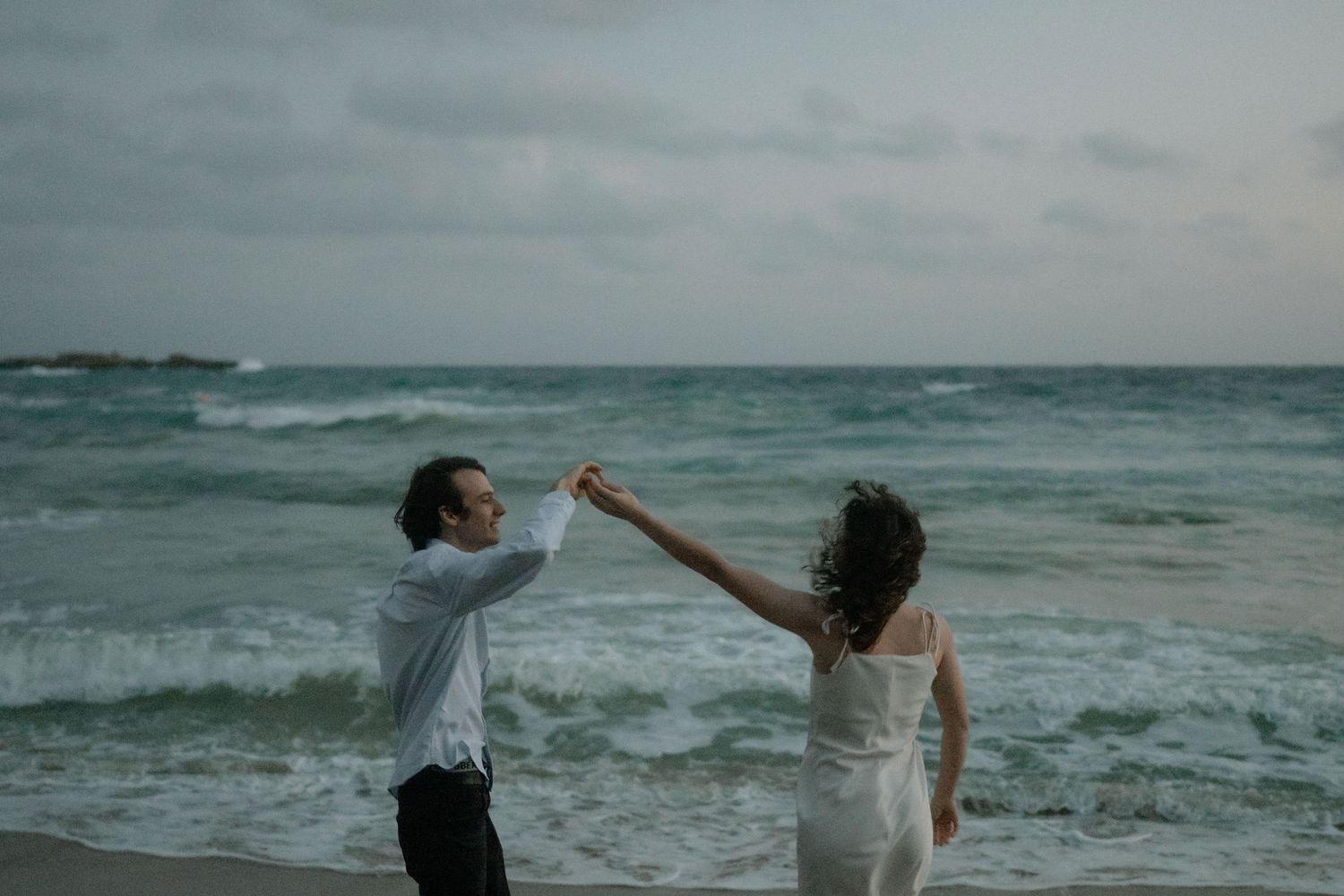 Couple dancing romantically on a beach with turquoise ocean waves and dramatic cloudy skies.