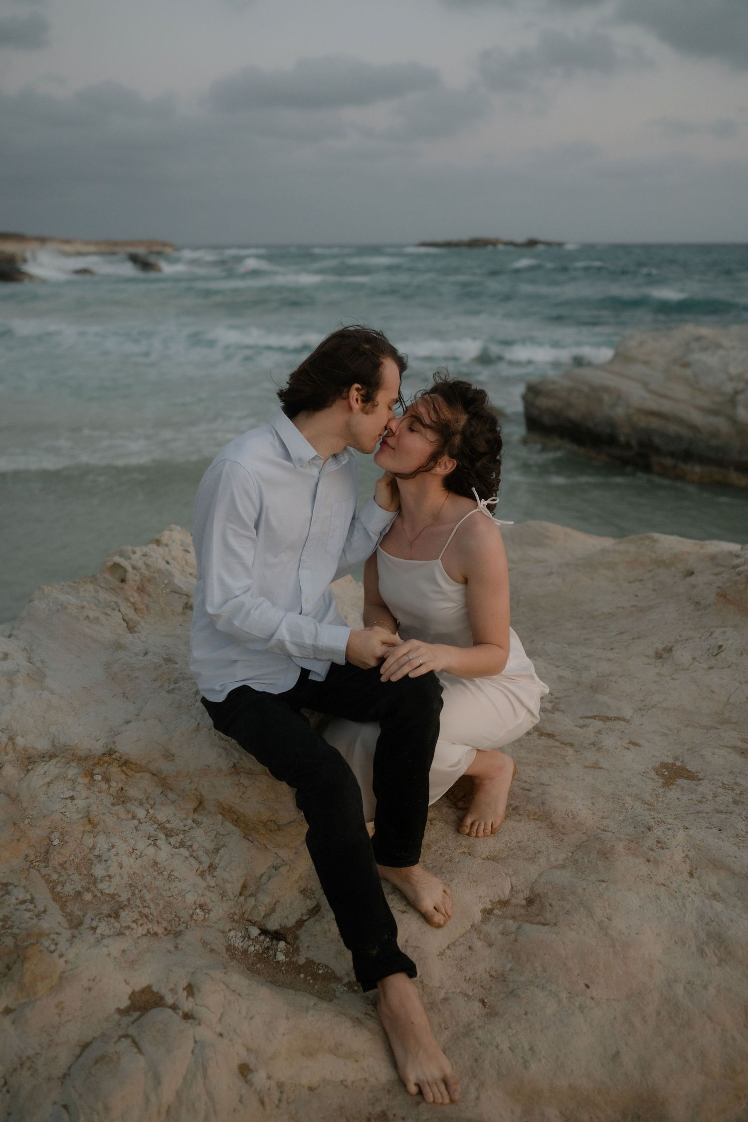 Couple sharing a romantic kiss on rocky seaside cliffs with turquoise ocean waves in the background.