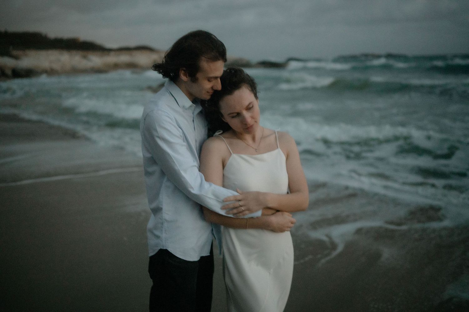 Romantic couple embracing on a moody beach, waves crashing behind them, woman in white dress, man in blue shirt.