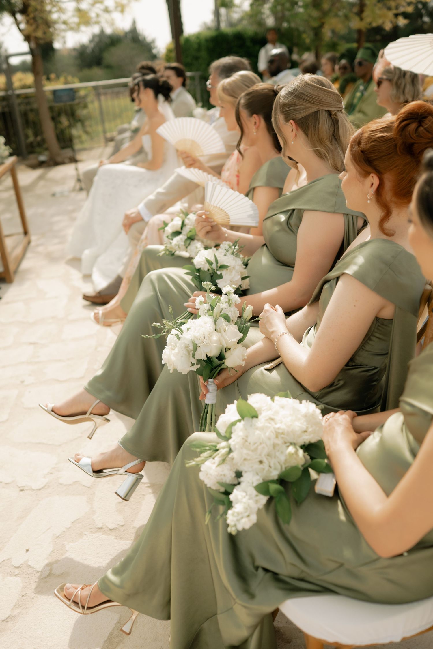 Bridesmaids in sage green dresses holding white bouquets seated at an outdoor wedding ceremony.