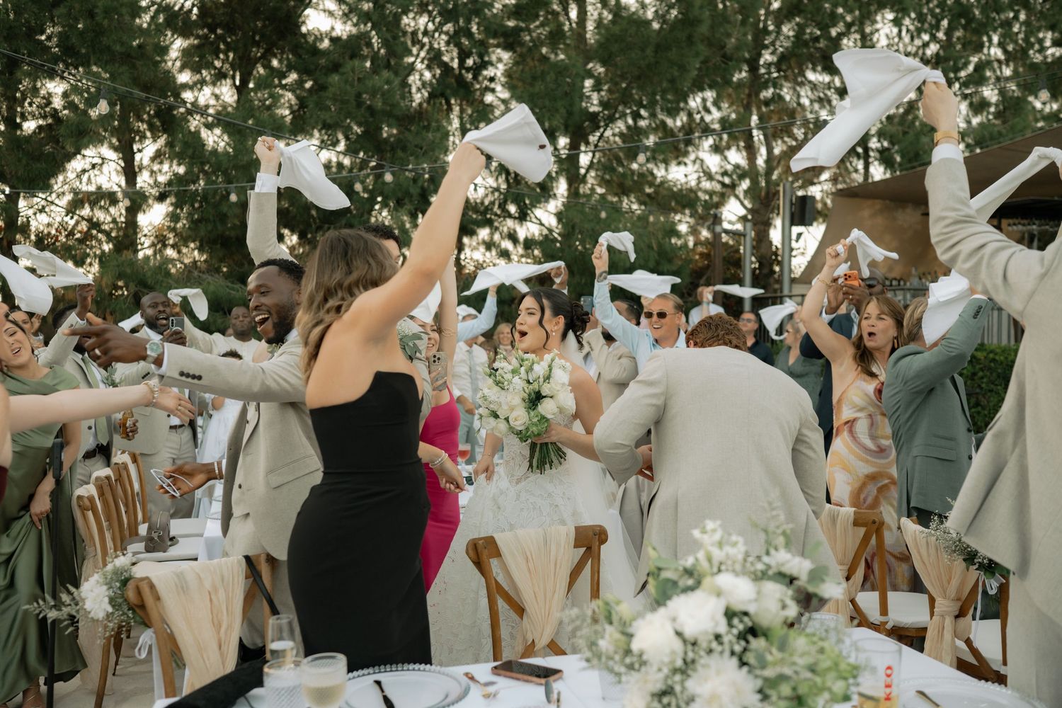 Wedding guests wave white napkins celebrating bride and groom at outdoor reception surrounded by lush green trees.