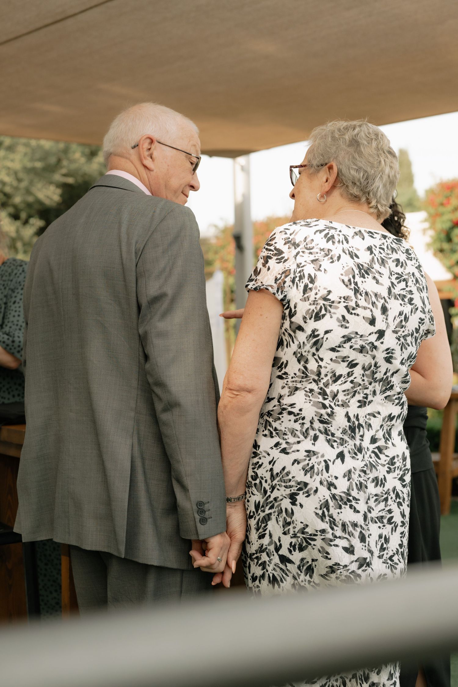 Elderly couple holding hands at outdoor event, man in gray suit and woman in floral dress, viewed from behind.