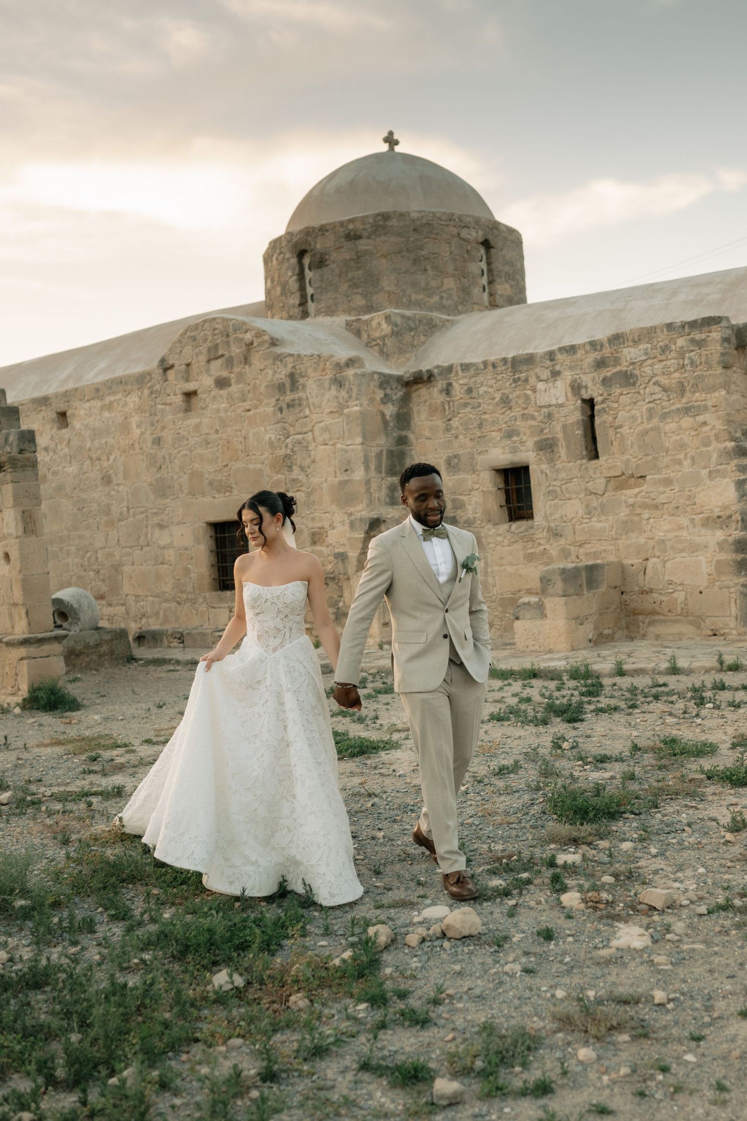 Bride and groom walk hand-in-hand before a historic stone church at sunset during their outdoor wedding.