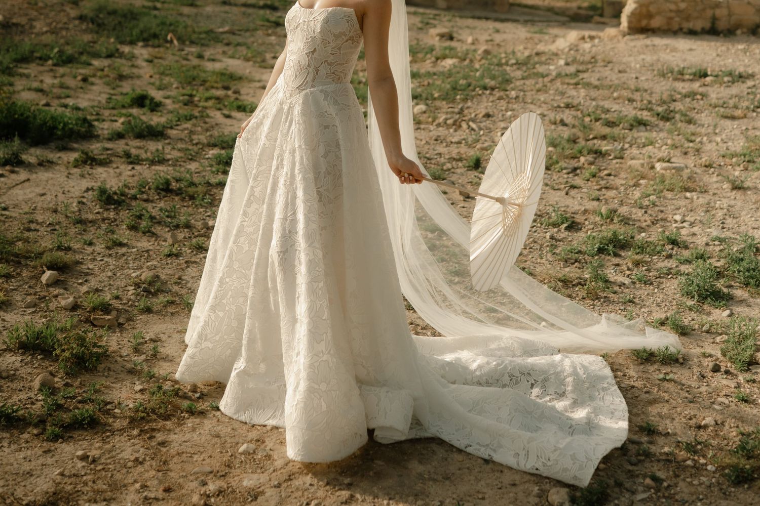 Bride in a flowing white lace wedding gown with a long train, standing outdoors on sandy ground.