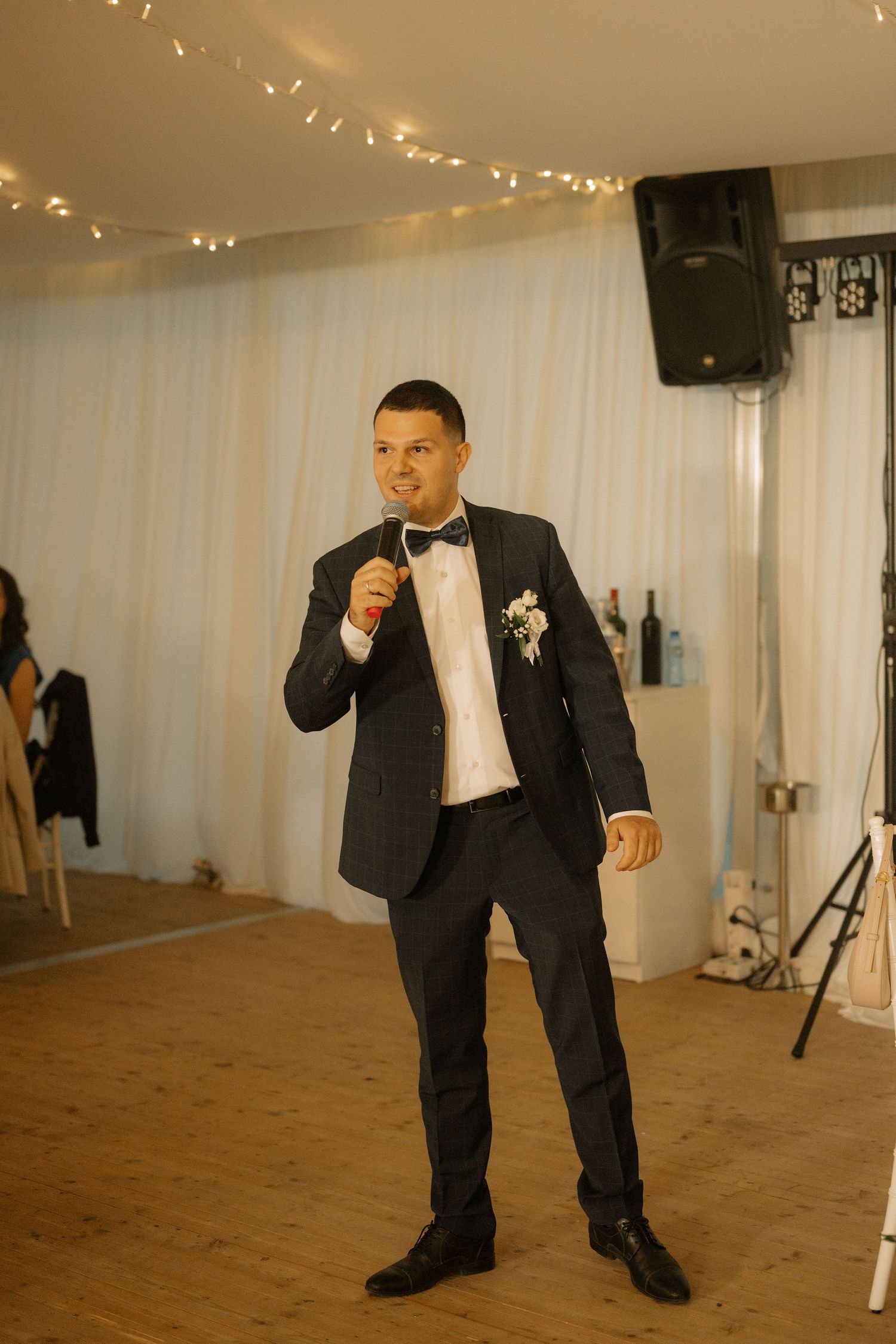 Groom in black tuxedo with bow tie and boutonniere speaking into microphone at wedding reception.