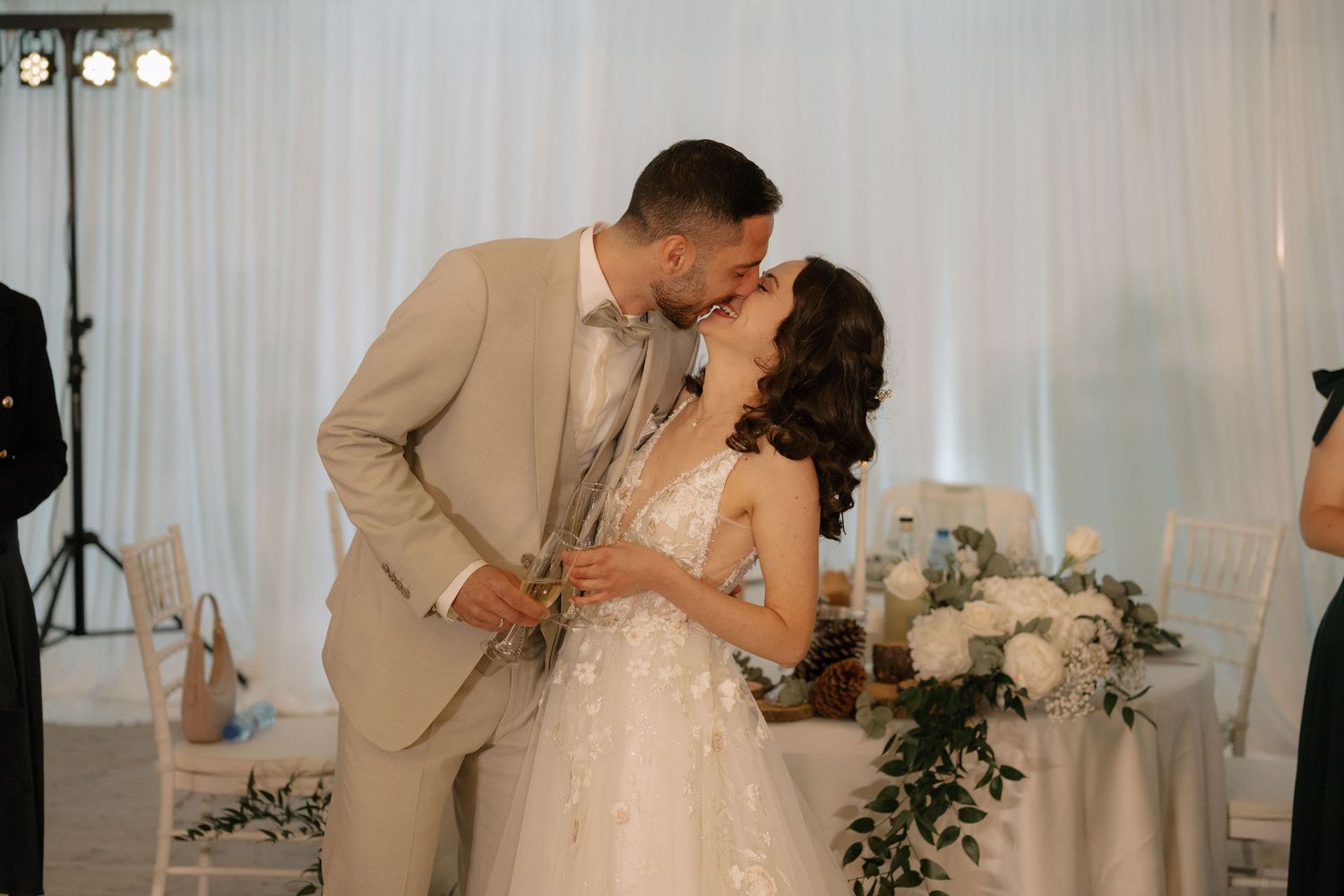 A bride and groom share a kiss at their elegant wedding reception with white floral arrangements and draped decor.