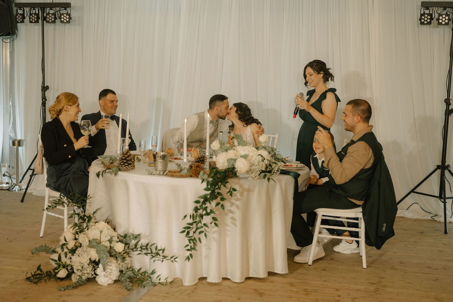 Wedding party toasts newlyweds at elegantly decorated sweetheart table with white florals and greenery.
