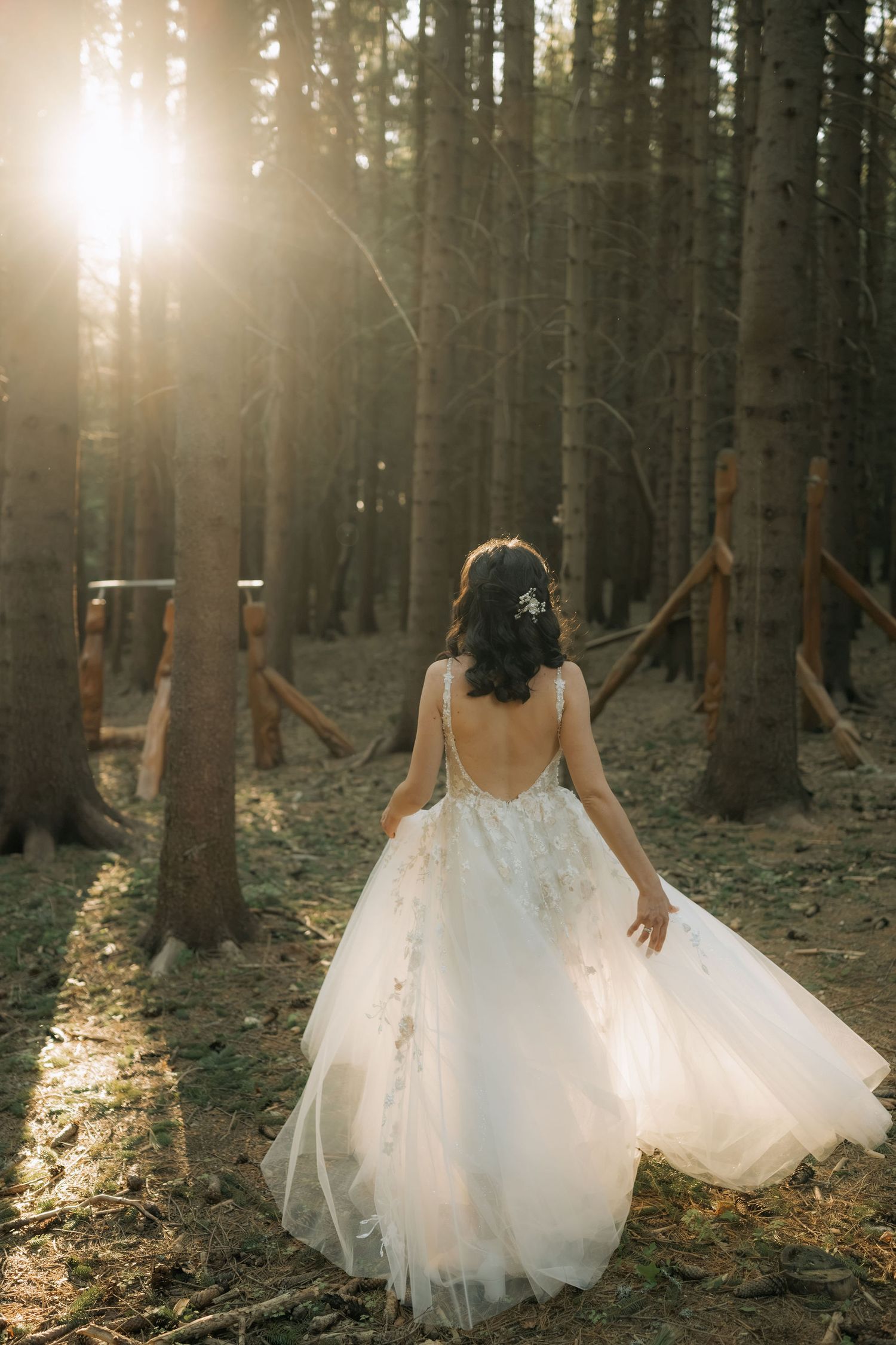 Bride in white backless floral gown walks through sunlit forest, hair adorned with flowers, dress trailing gracefully.