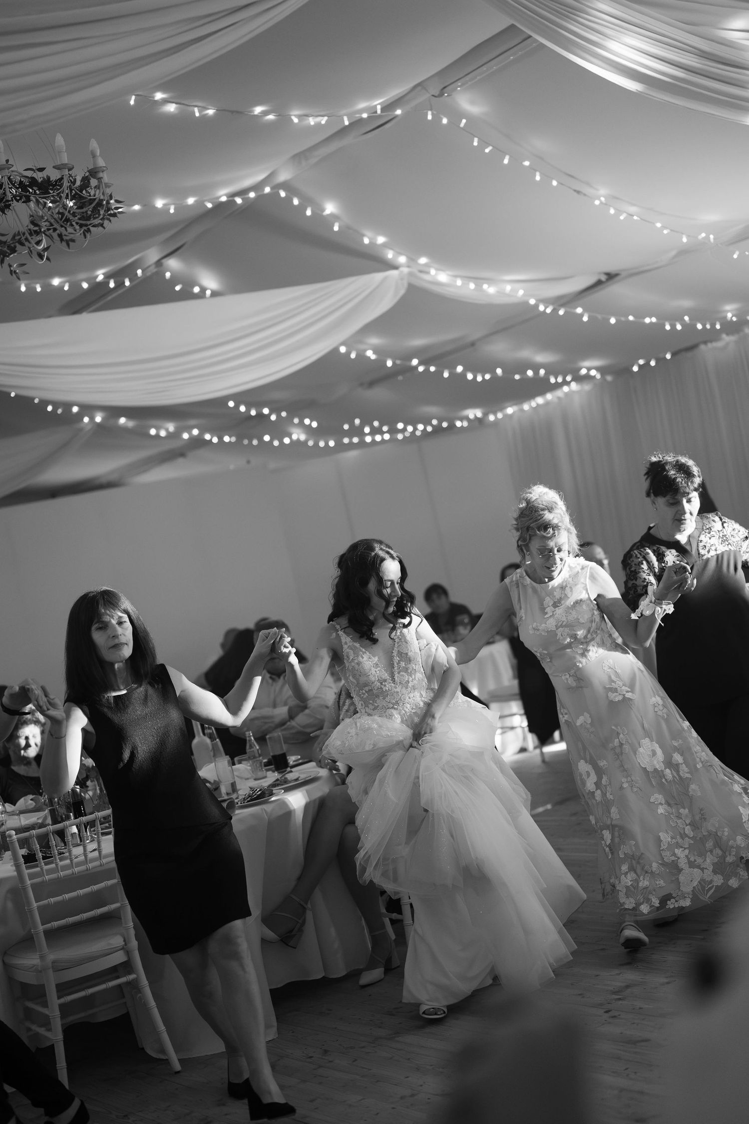 Black and white photo of a bride dancing joyfully with guests under fairy lights at an elegant wedding reception.