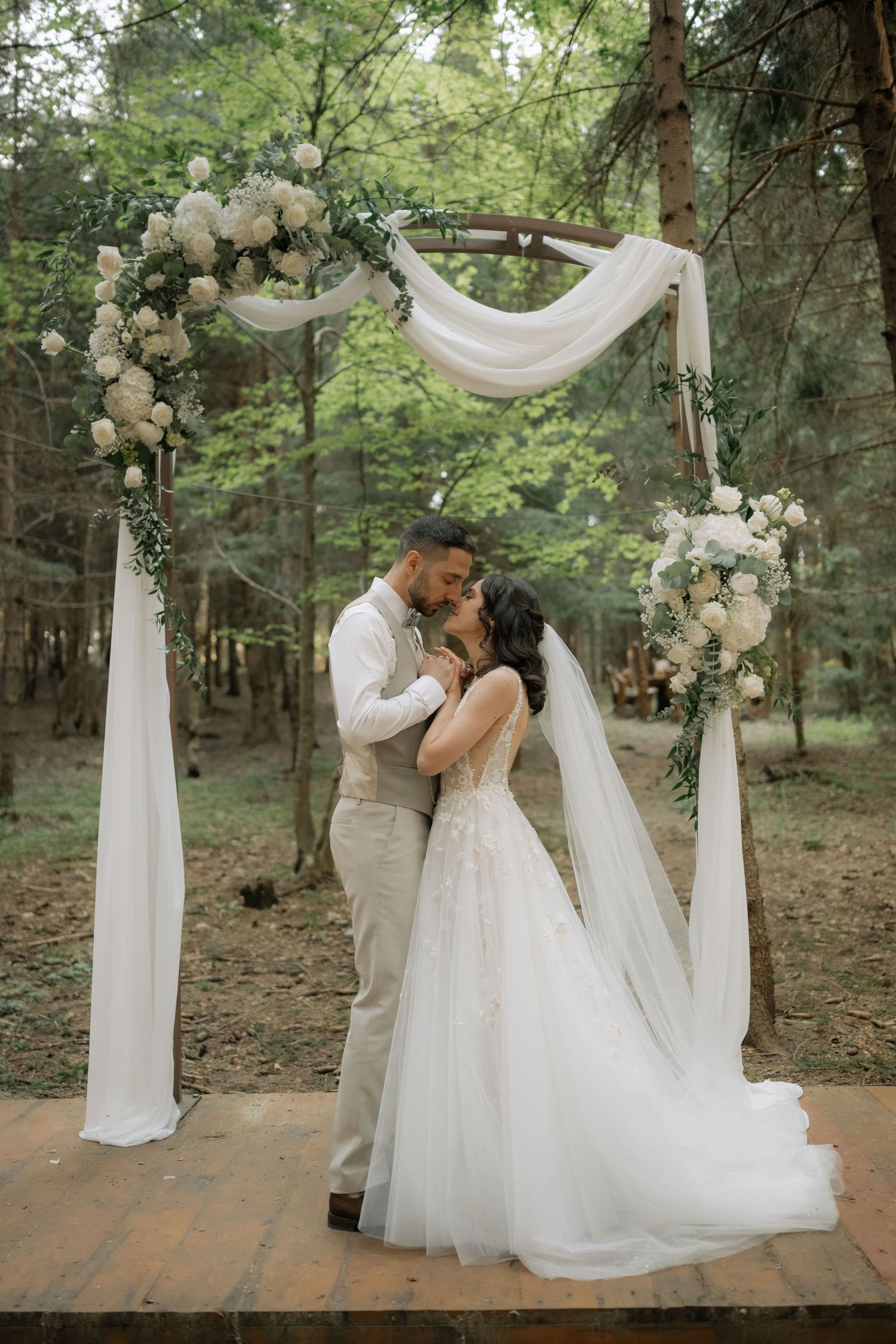 Bride and groom share a romantic kiss under a floral arch with white draping in a lush forest wedding ceremony.