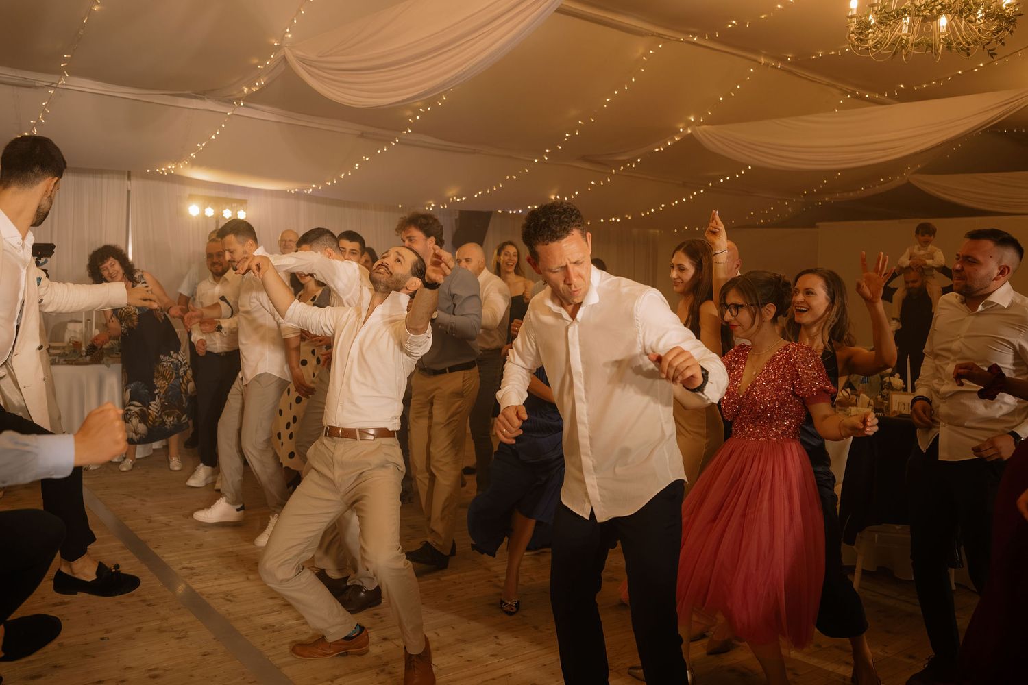 Guests dancing energetically at a wedding reception in a decorated tent venue with string lights.