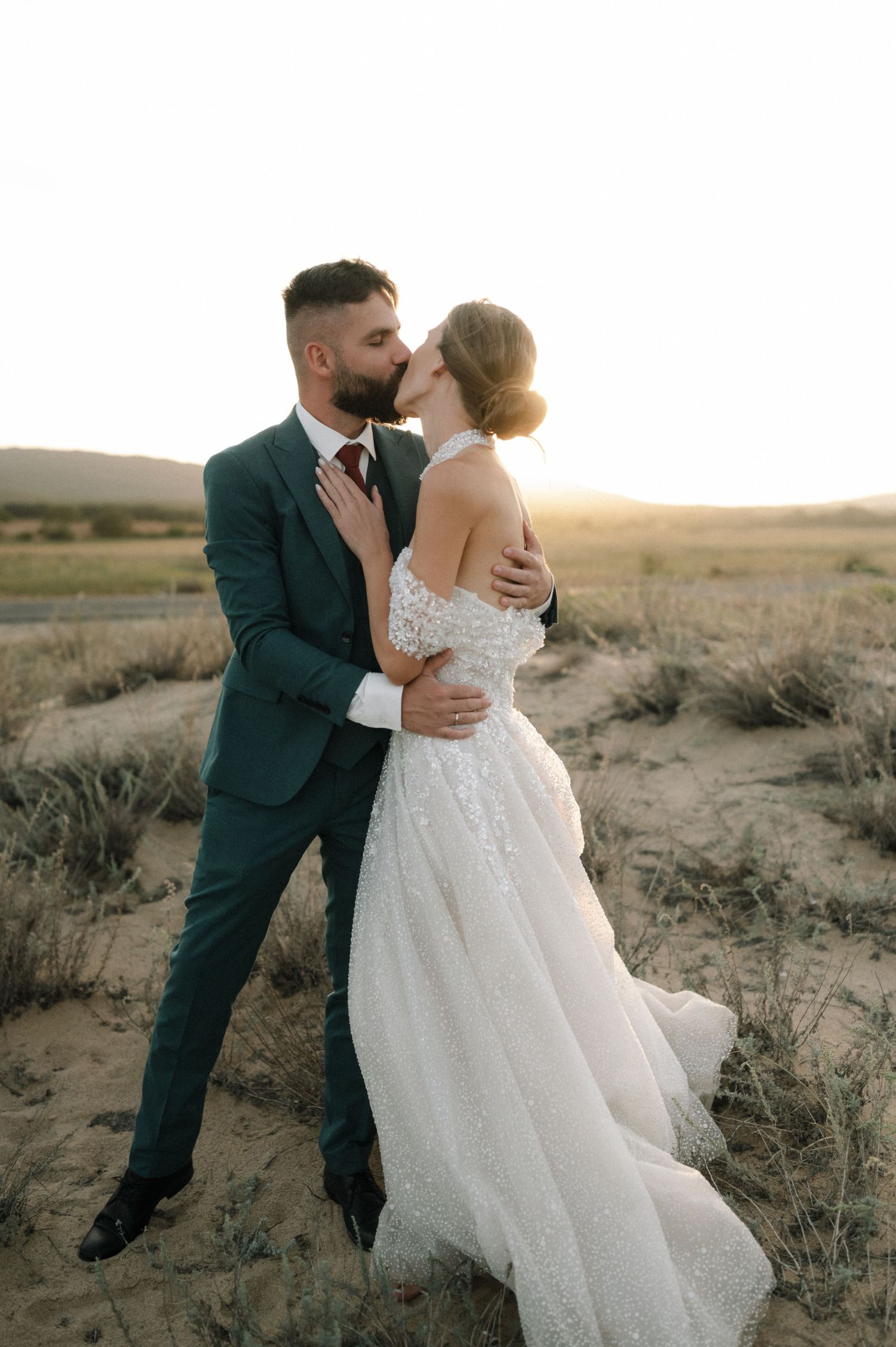 A bride and groom share a romantic kiss at sunset in a desert landscape, she in a flowing white gown, he in a dark green suit.
