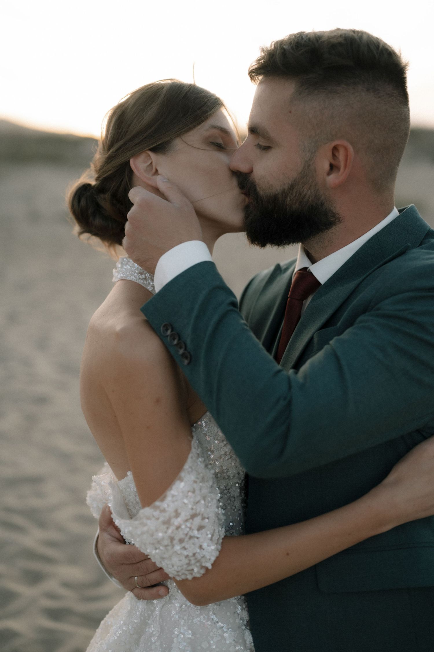 A bride and groom share a romantic kiss on a sandy beach at sunset, dressed in a white gown and teal suit.
