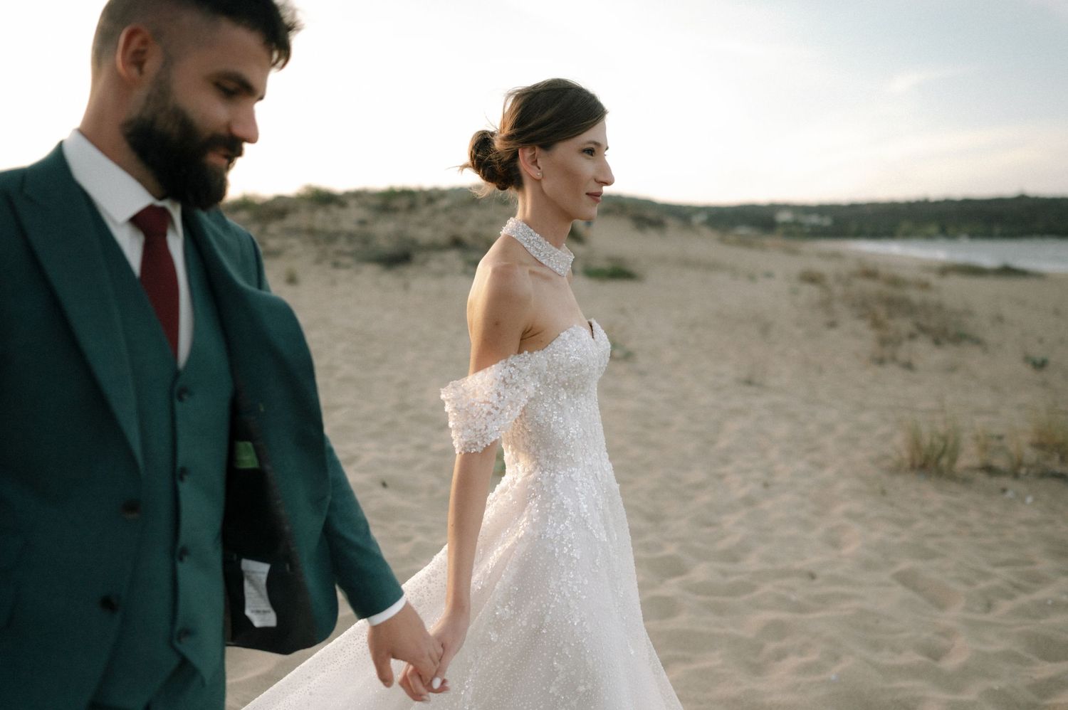 Bride and groom walking hand in hand on sandy beach, woman in lace off-shoulder gown, man in green suit.