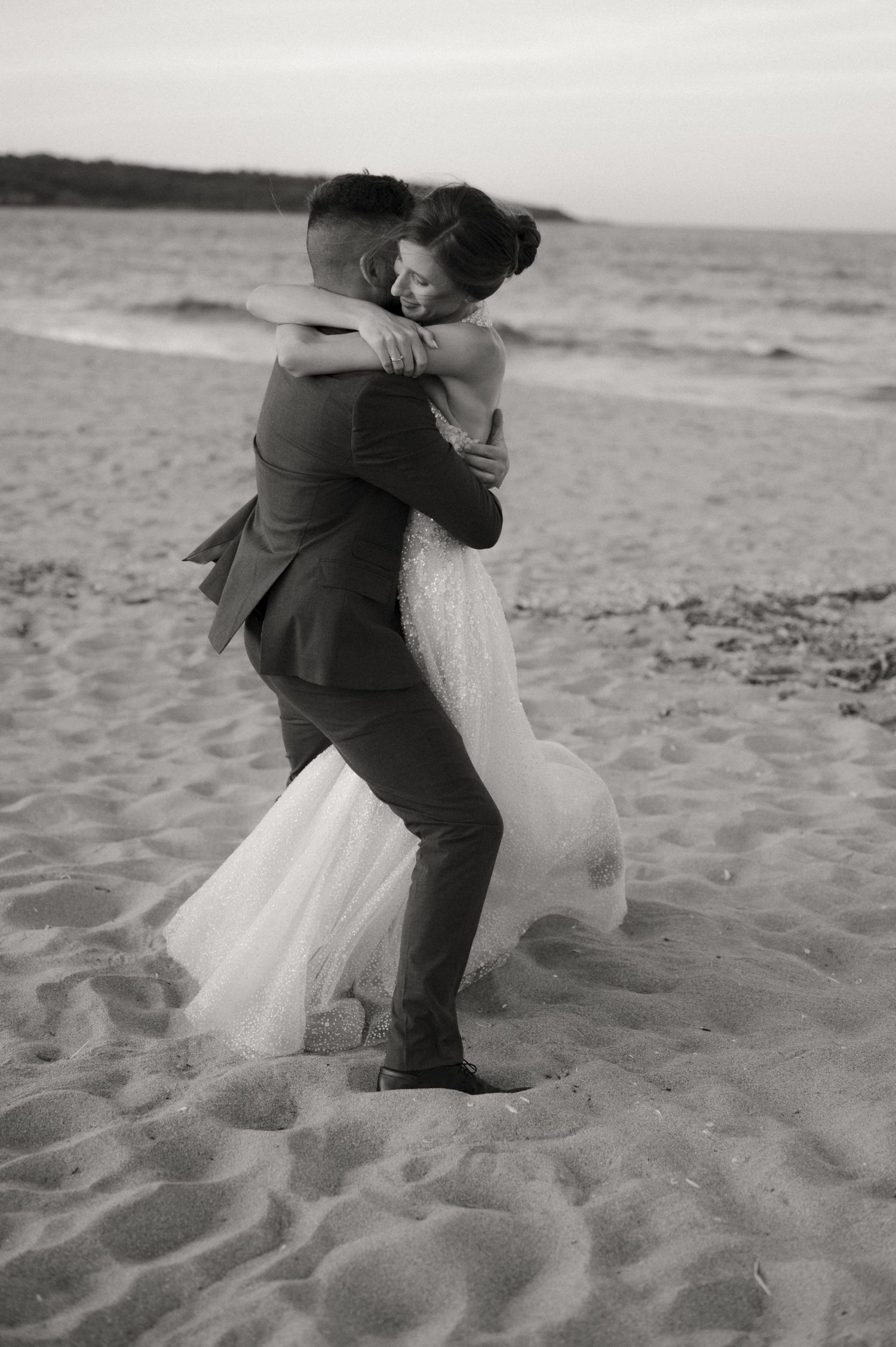 Black and white photo of a groom lifting his bride in a wedding dress on a sandy beach.