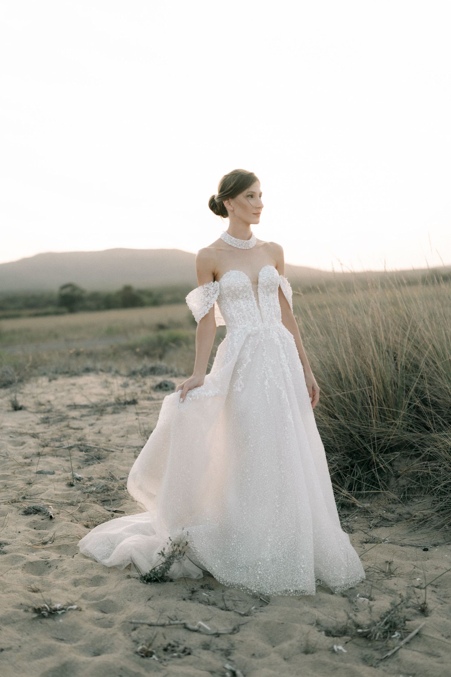 Bride in a sparkling off-shoulder ball gown with floral lace bodice stands on sandy dunes at golden hour.