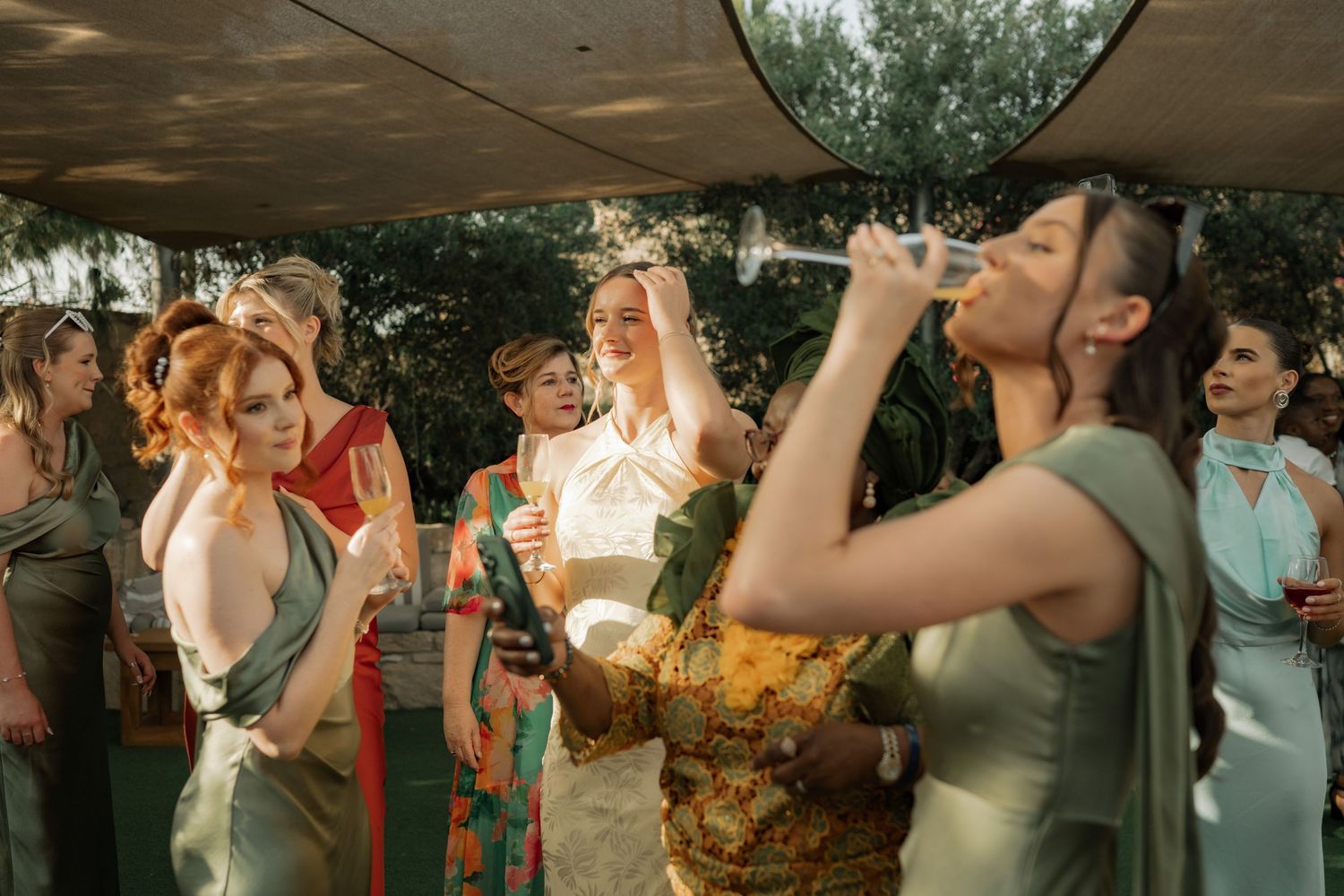 Women in elegant dresses celebrate at an outdoor wedding reception, with one guest taking a celebratory drink.