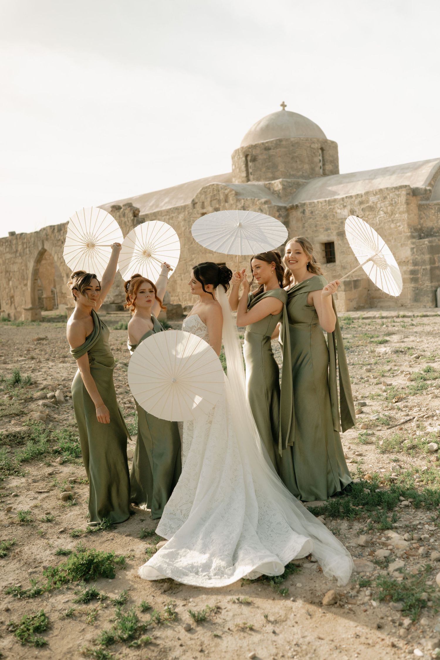 Bride in white gown with bridesmaids in olive green dresses holding white parasols near ancient stone church ruins.
