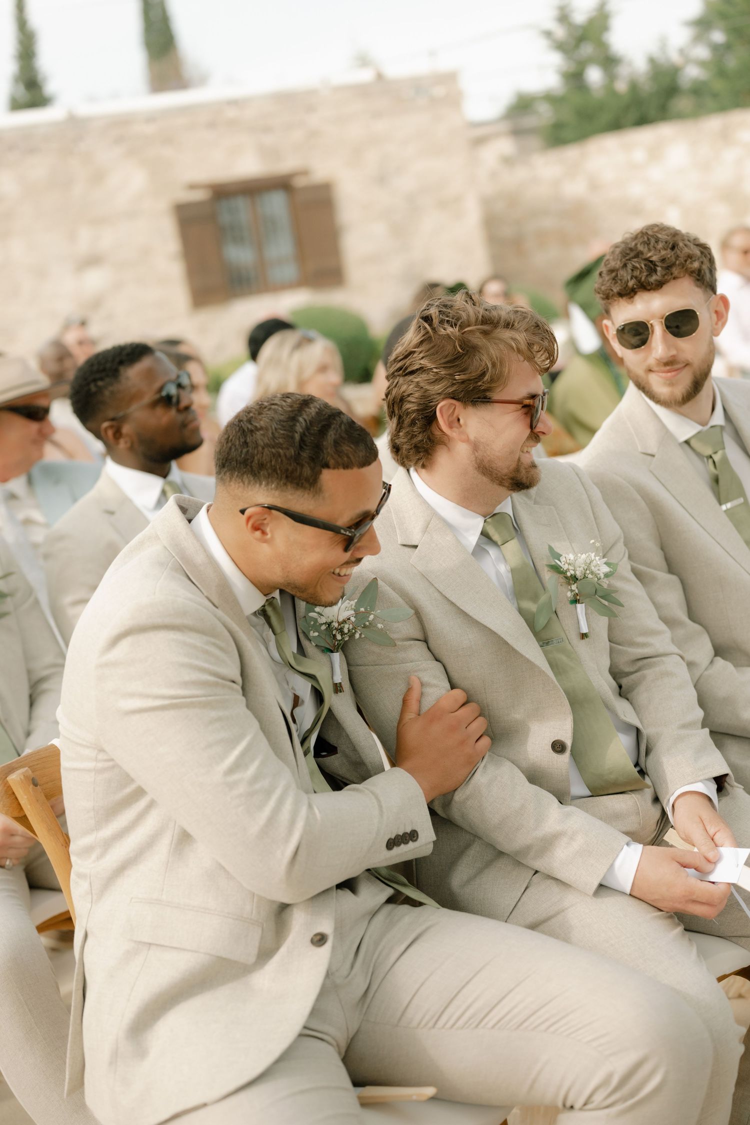 Men in beige suits with boutonnieres sit at an outdoor wedding ceremony, smiling and enjoying the celebration.