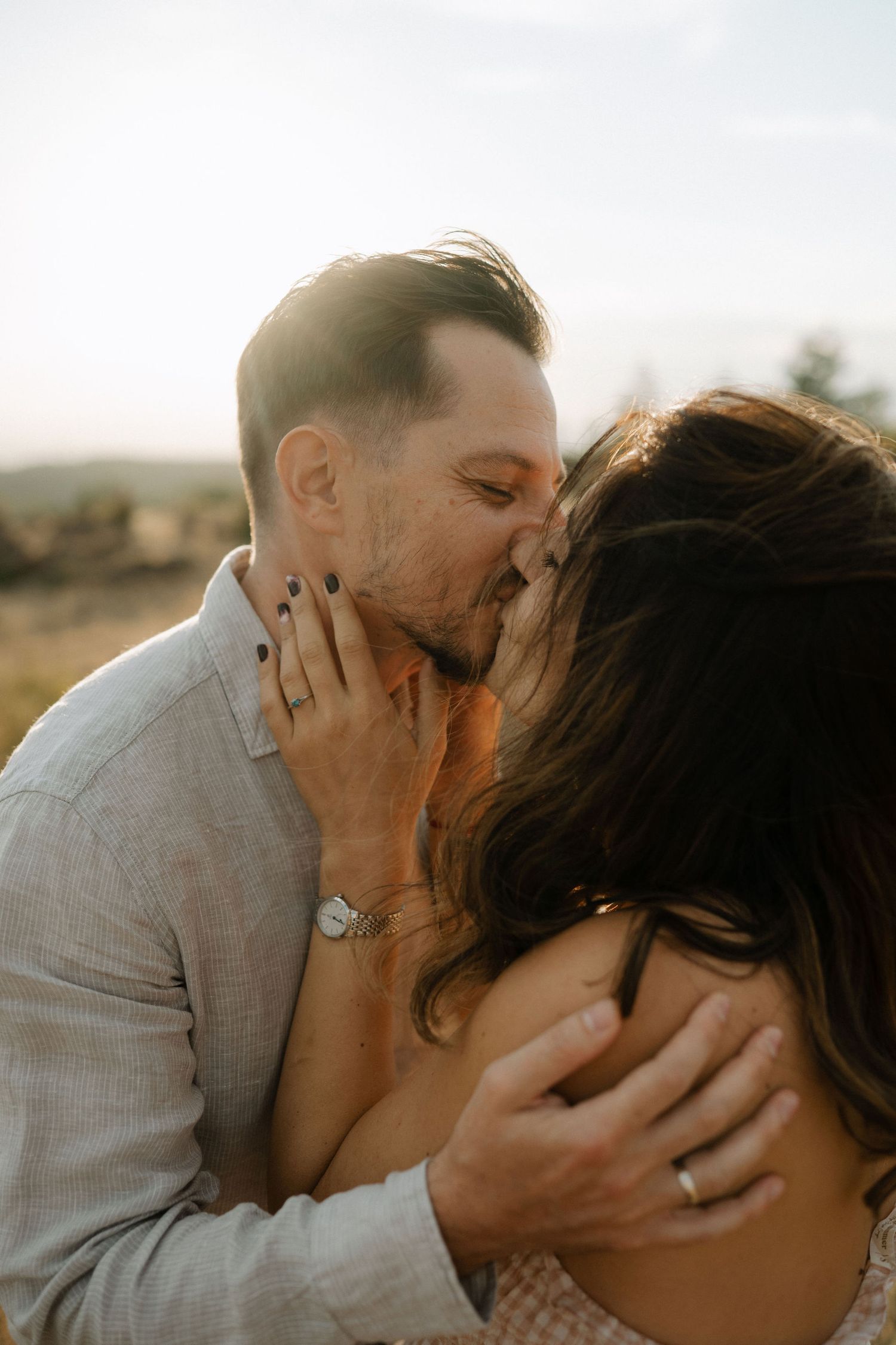 Romantic couple shares intimate kisses in warm sunlight wearing light colored clothing against desert landscape.