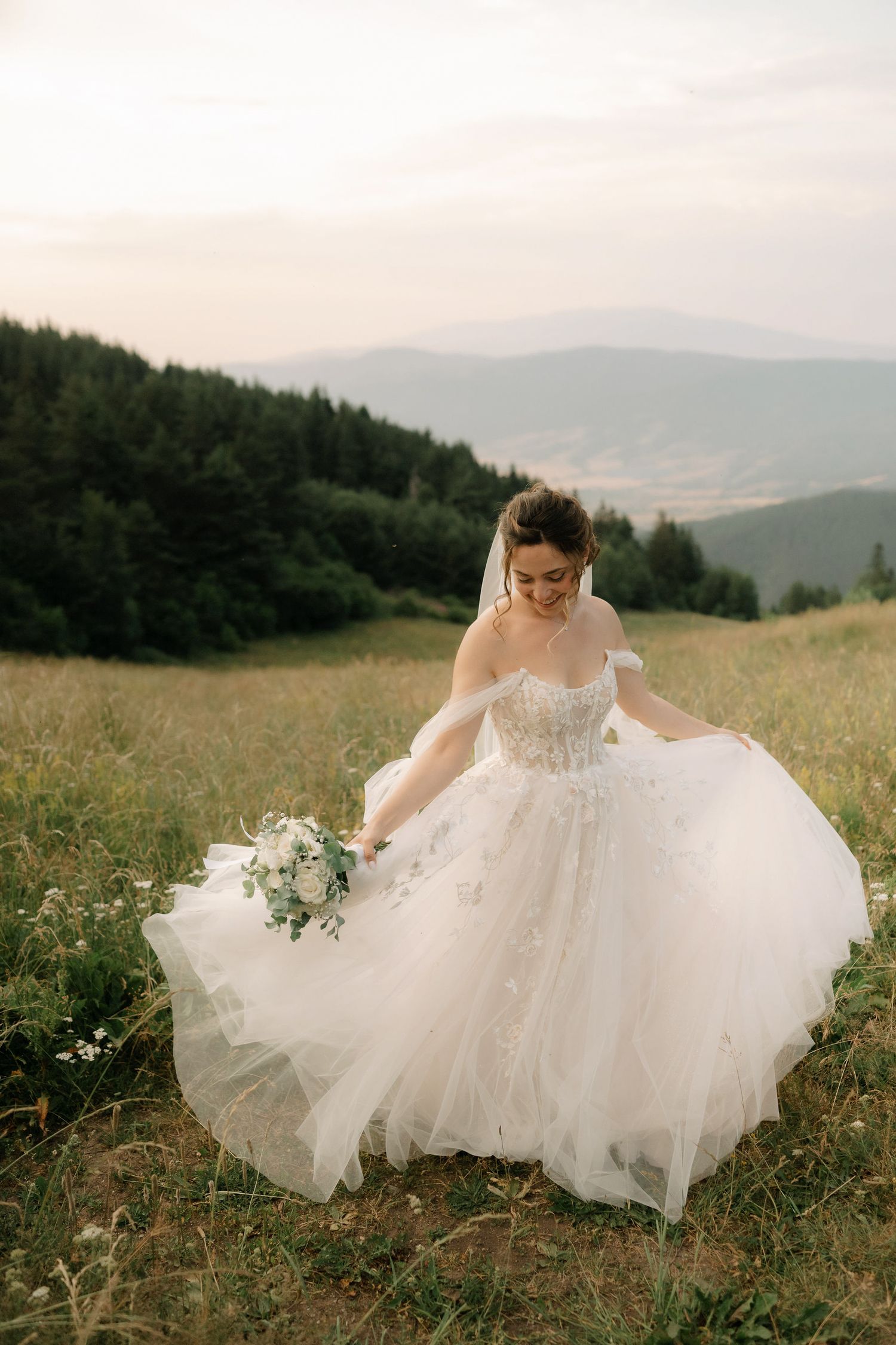 A flowing white wedding dress twirls gracefully in a mountain meadow with wildflowers and evergreen forests in the background.