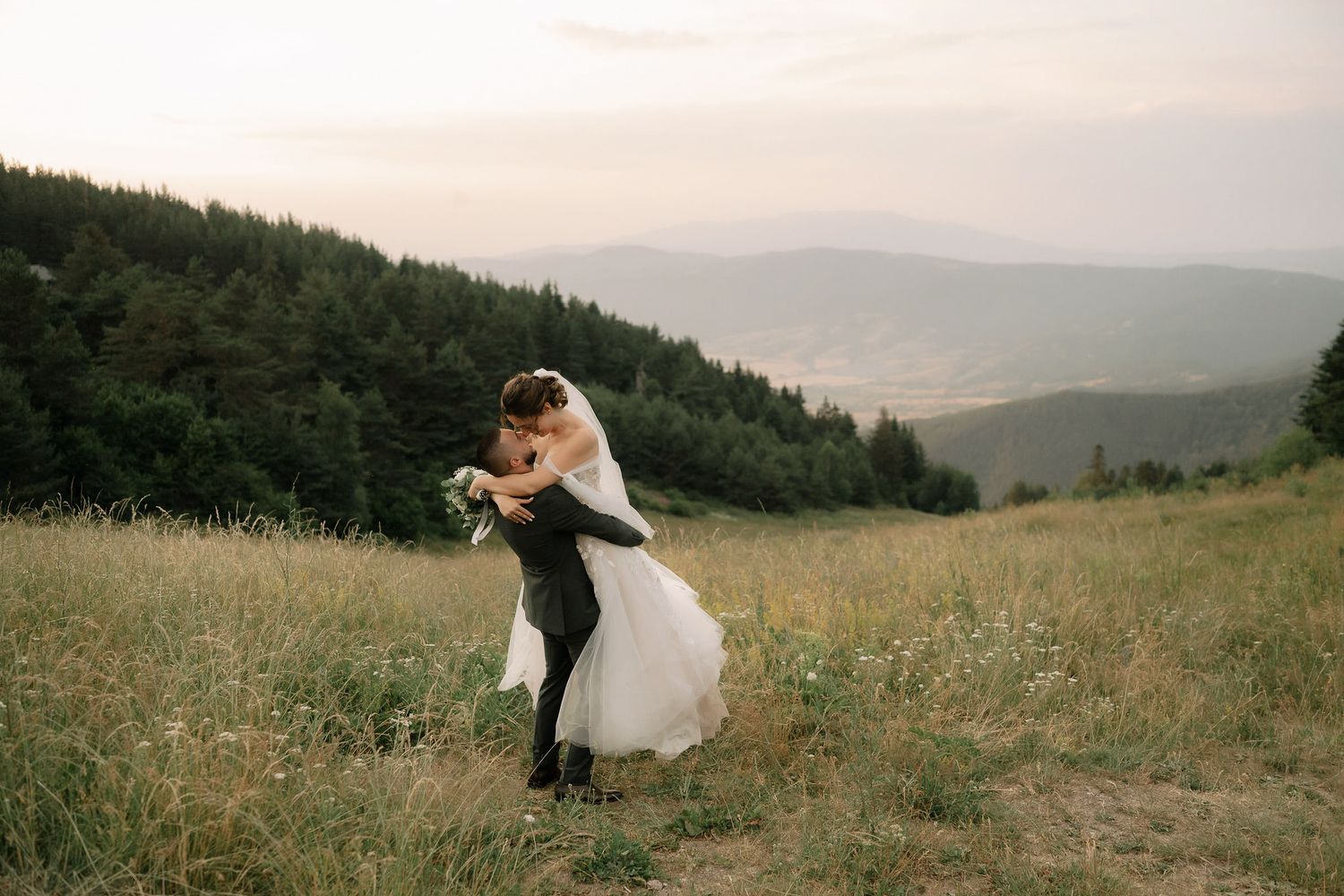 A couple embraces on a scenic mountaintop meadow at sunset with rolling hills and forest in the background.