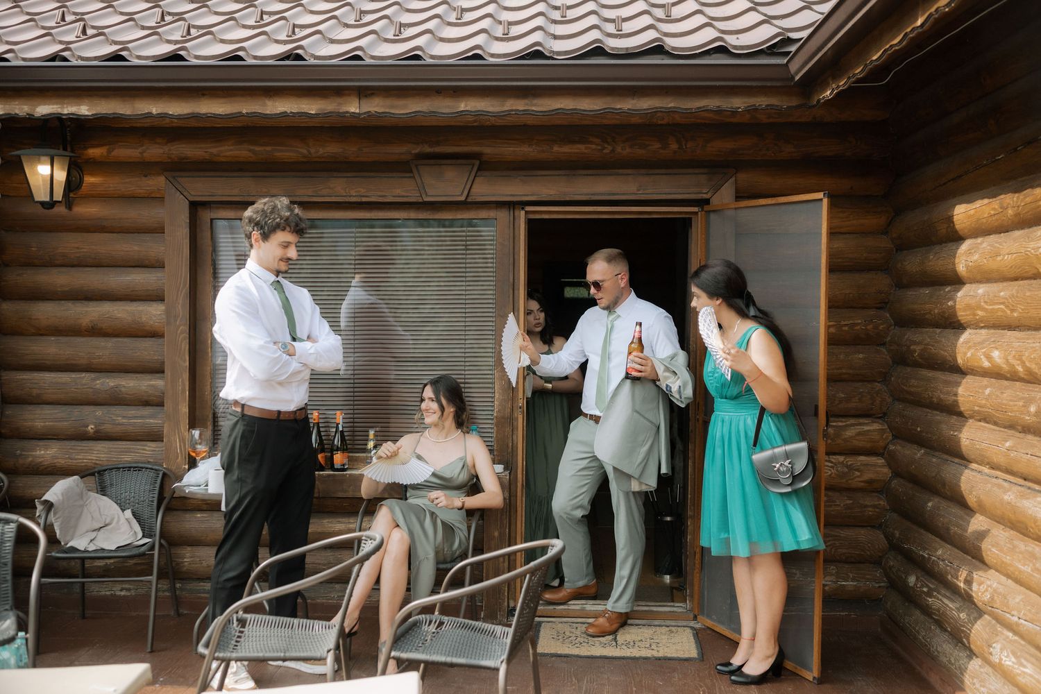 People gather on a rustic log cabin porch during a social event on a summer day.