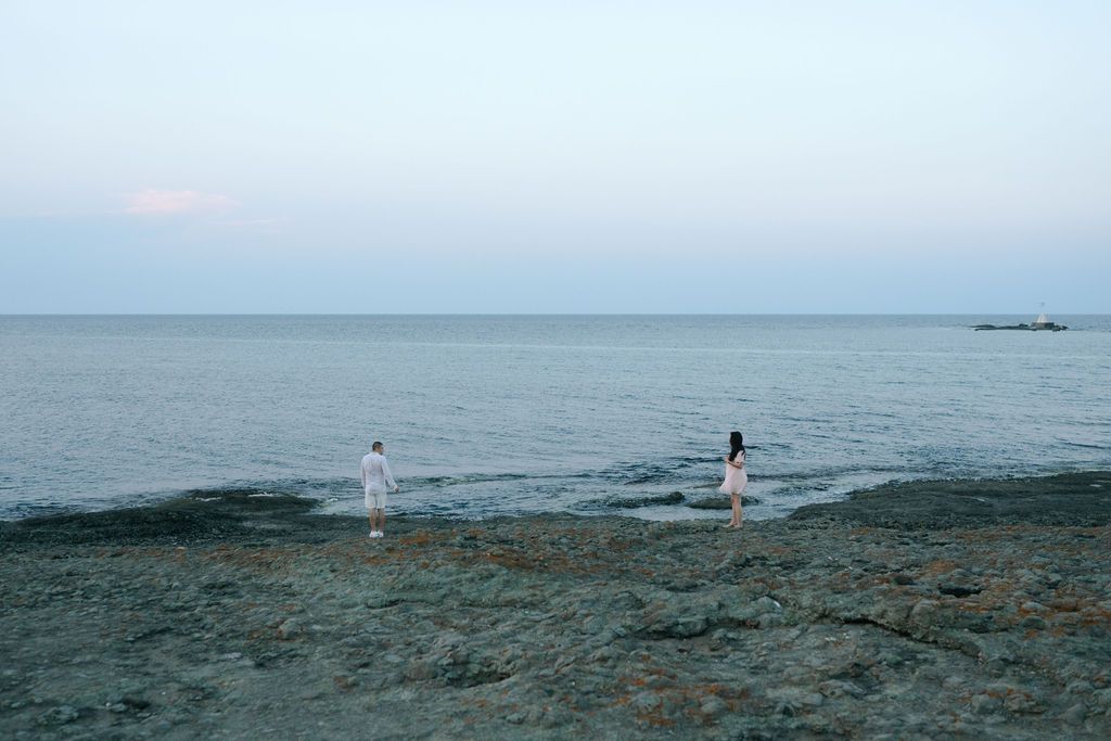 A person stands at the edge of a rocky beach looking out towards the calm ocean horizon.