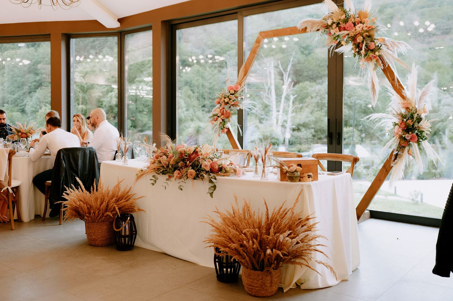 Modern wedding reception setup with hexagonal floral arch, dried pampas grass decor and white tablecloths in sunlit venue.