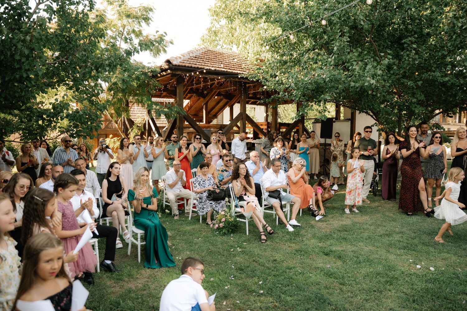 Outdoor wedding guests seated on wooden chairs in a garden setting during a summer ceremony.