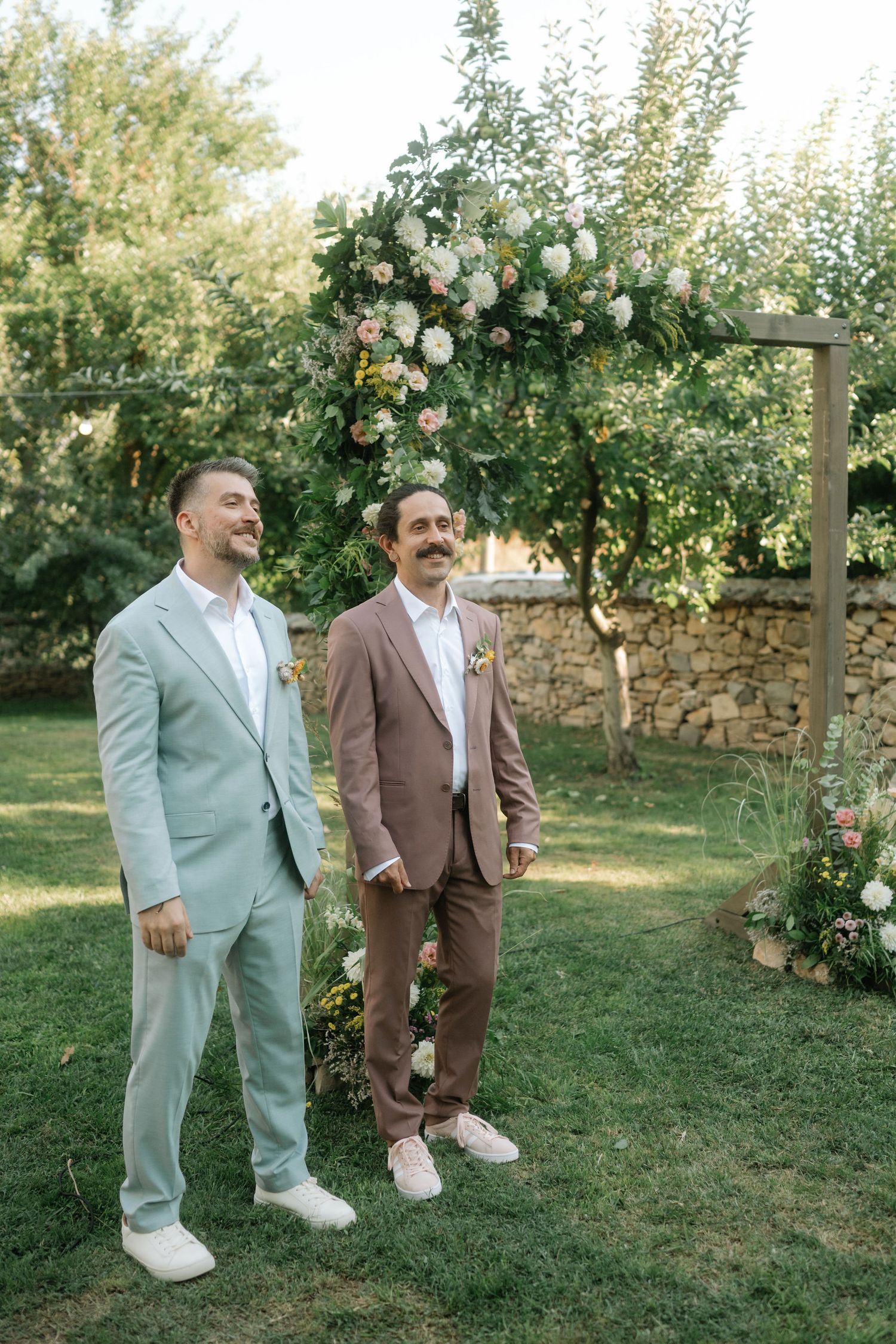 Two people in contrasting suits of mint green and brown stand together in a garden with blooming roses and a stone wall.