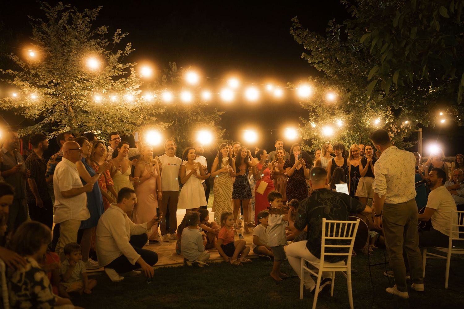 String lights illuminate an outdoor evening wedding reception with guests mingling and dining at tables.