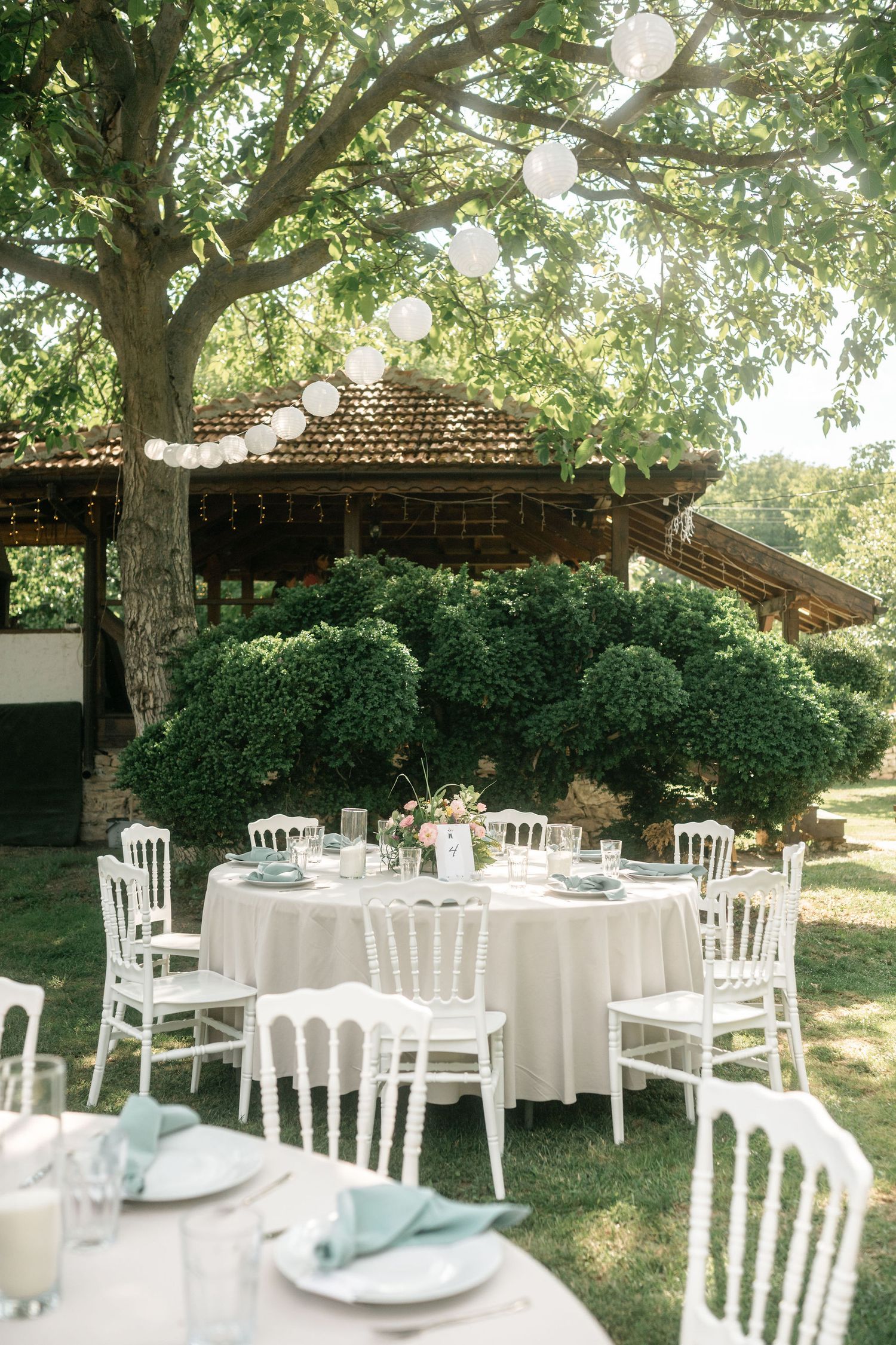 Elegant outdoor wedding reception setup with white round tables and chairs under a rustic wooden pavilion and shady tree.
