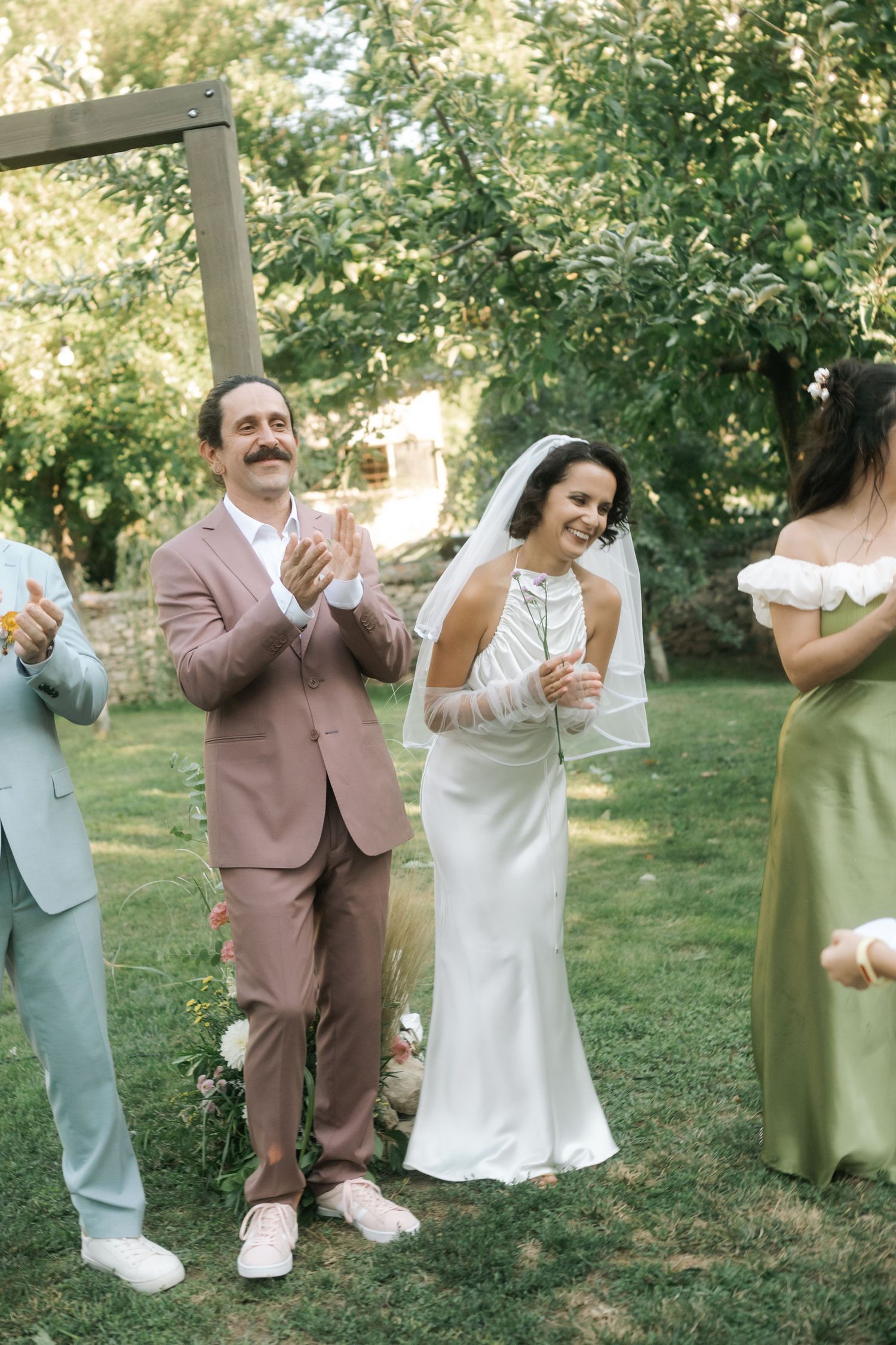 Wedding party members in pastel suits and dresses share a candid moment outdoors on a sunny day.