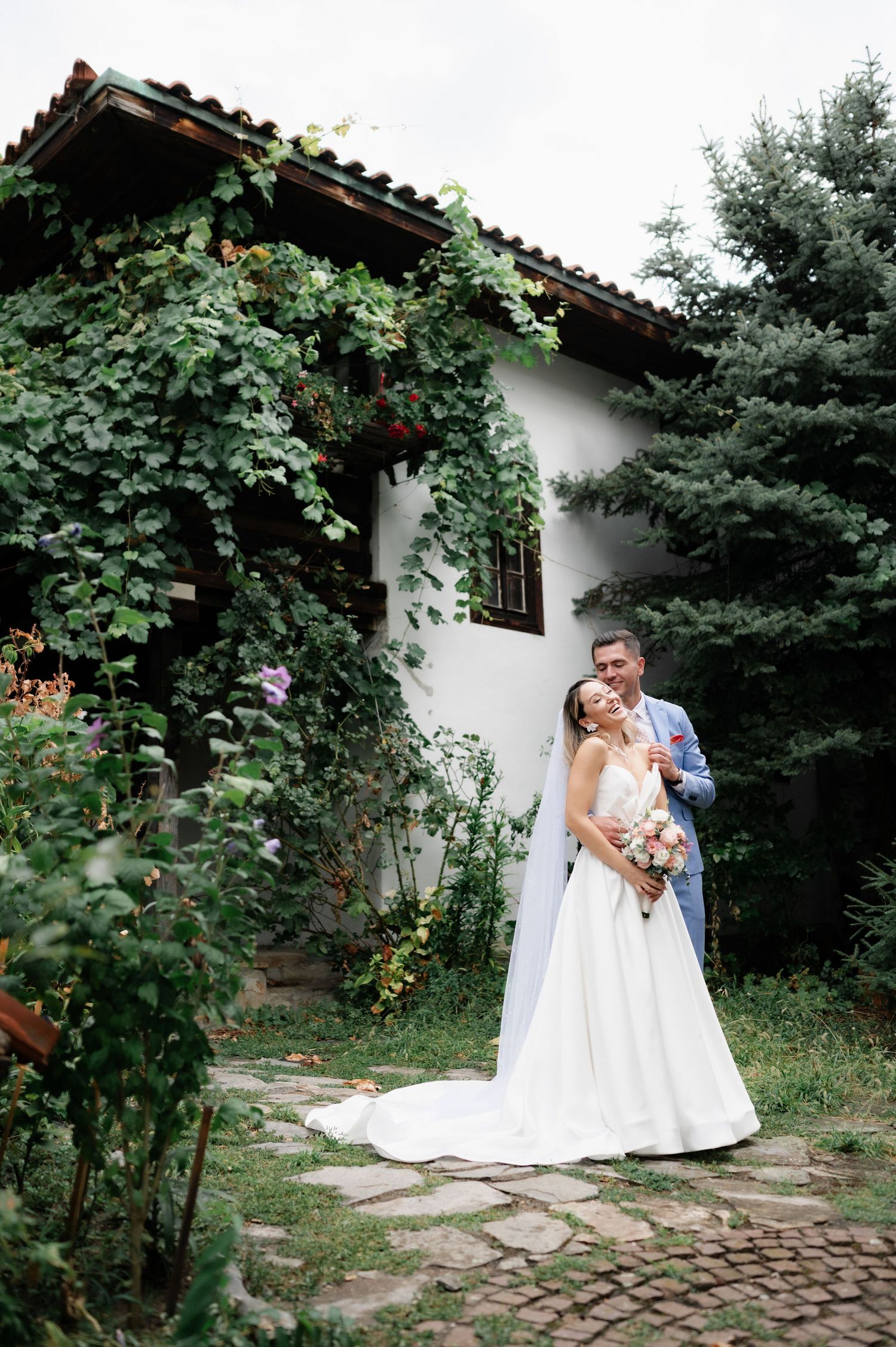 A person in a white wedding dress poses in a garden setting with stone pathways and lush greenery.