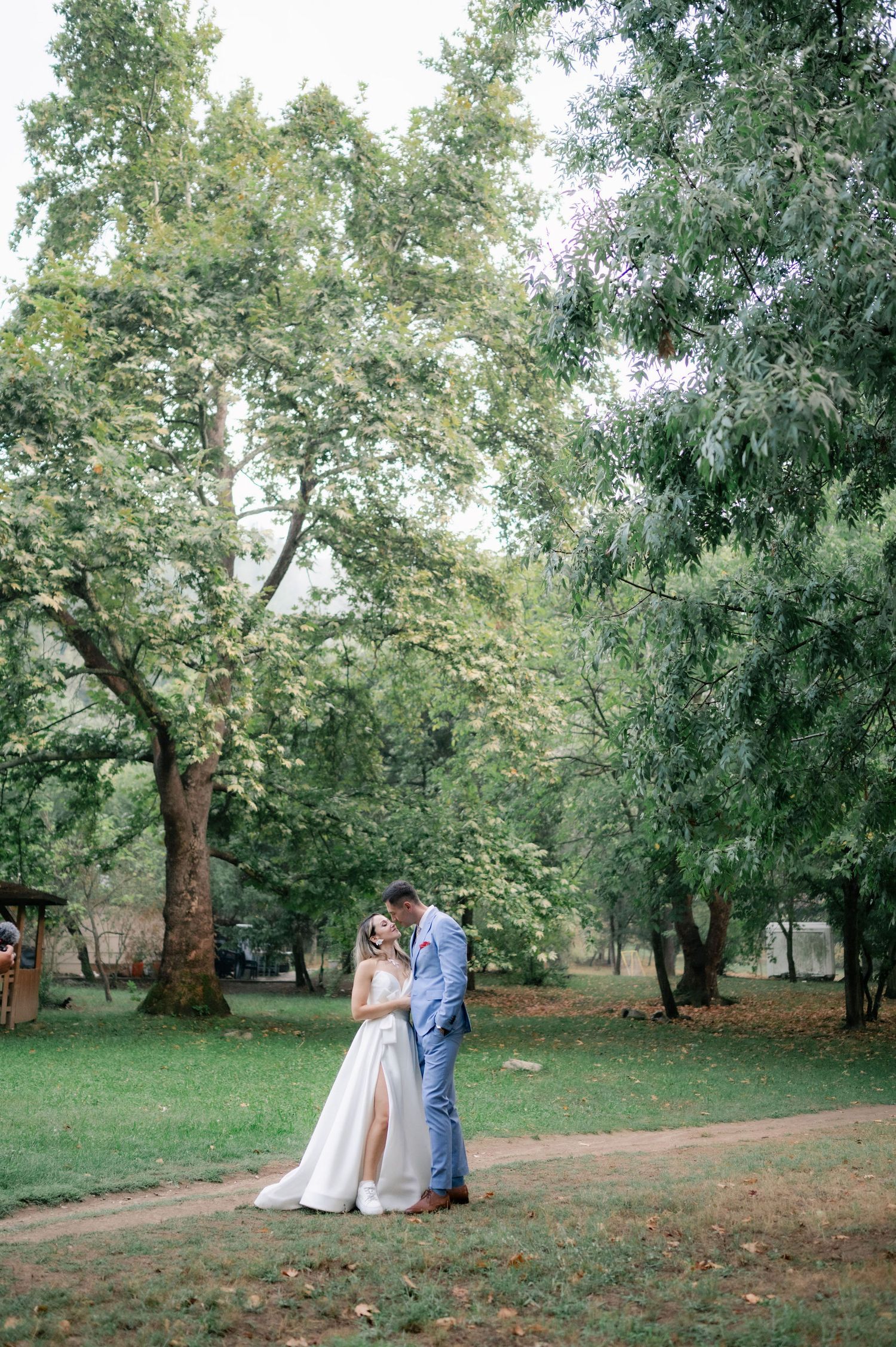 A couple in formal attire poses for romantic wedding photos under large shady trees in a peaceful park setting.