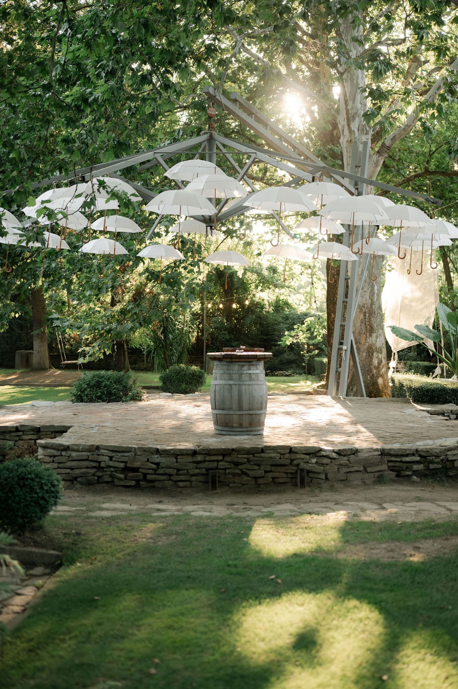 White umbrellas provide shade over rustic stone seating areas in a tranquil garden setting with dappled sunlight.