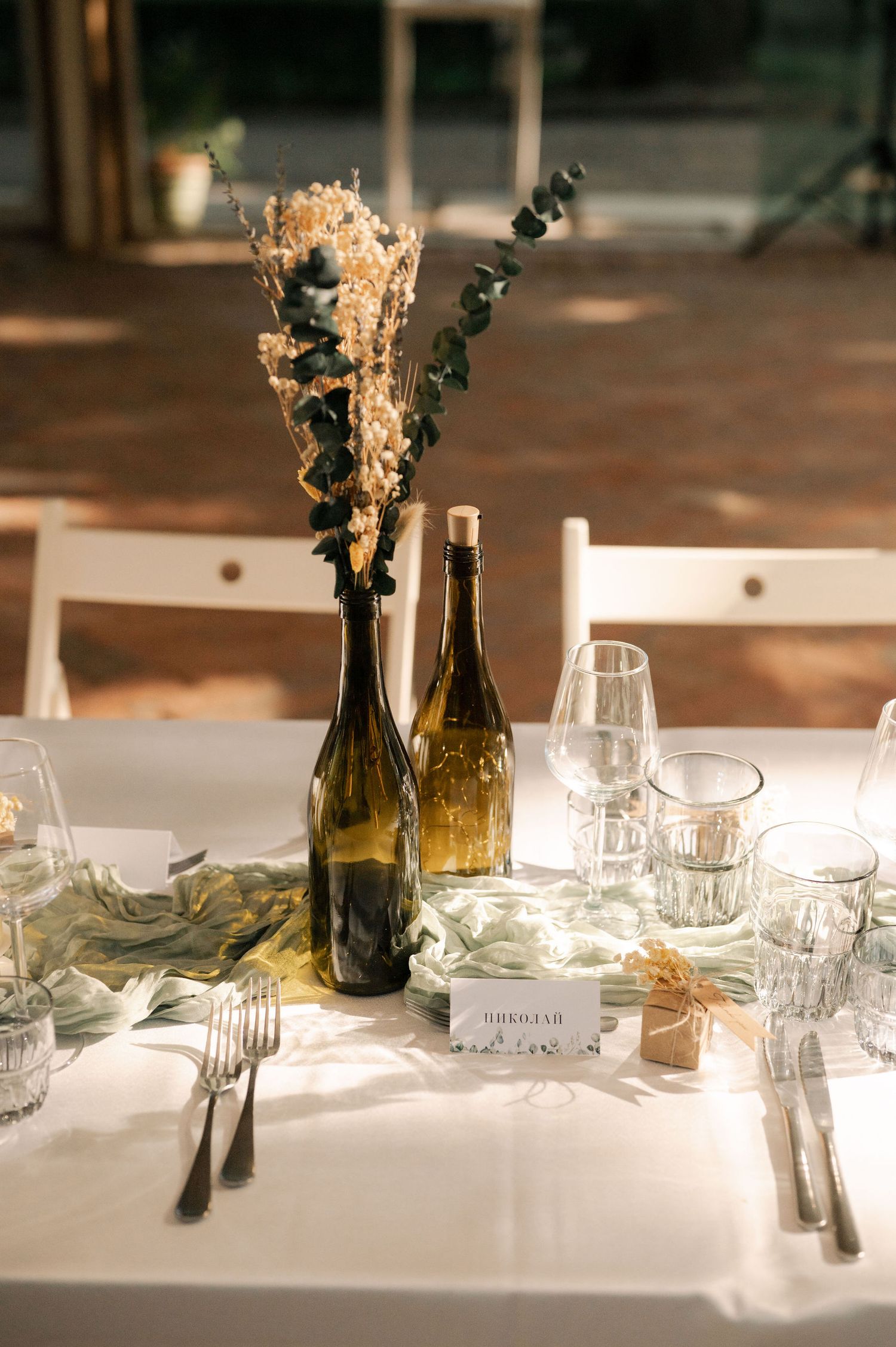 Wedding reception table settings with wine bottles, glassware, and dried flower centerpieces on white tablecloths.