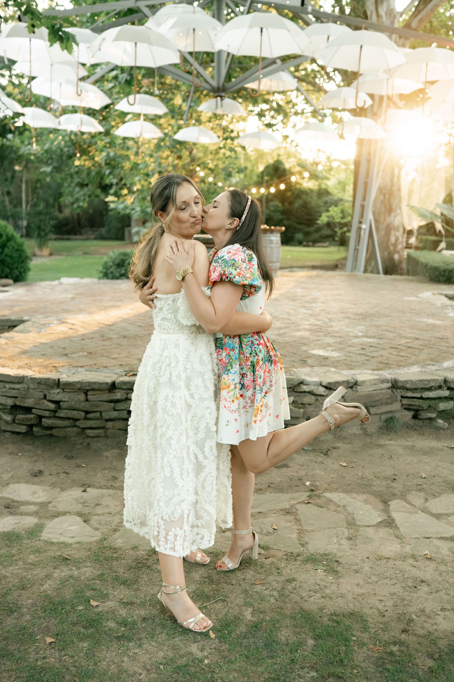 Two people in white and floral dresses share tender moments on a stone patio during a warm summer evening.