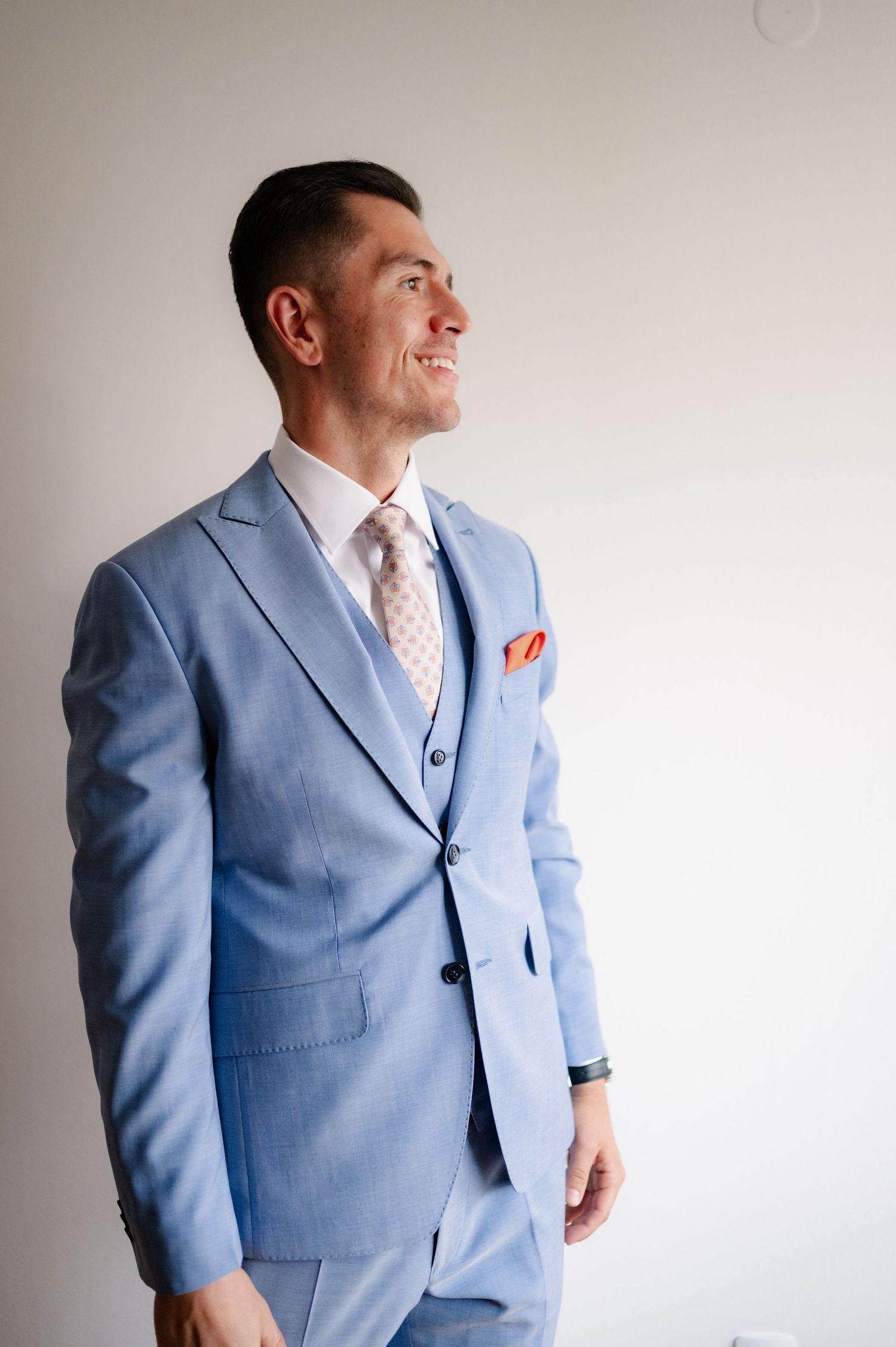 A man wearing a light blue three-piece suit and peach tie poses against a white wall in a series of shots.