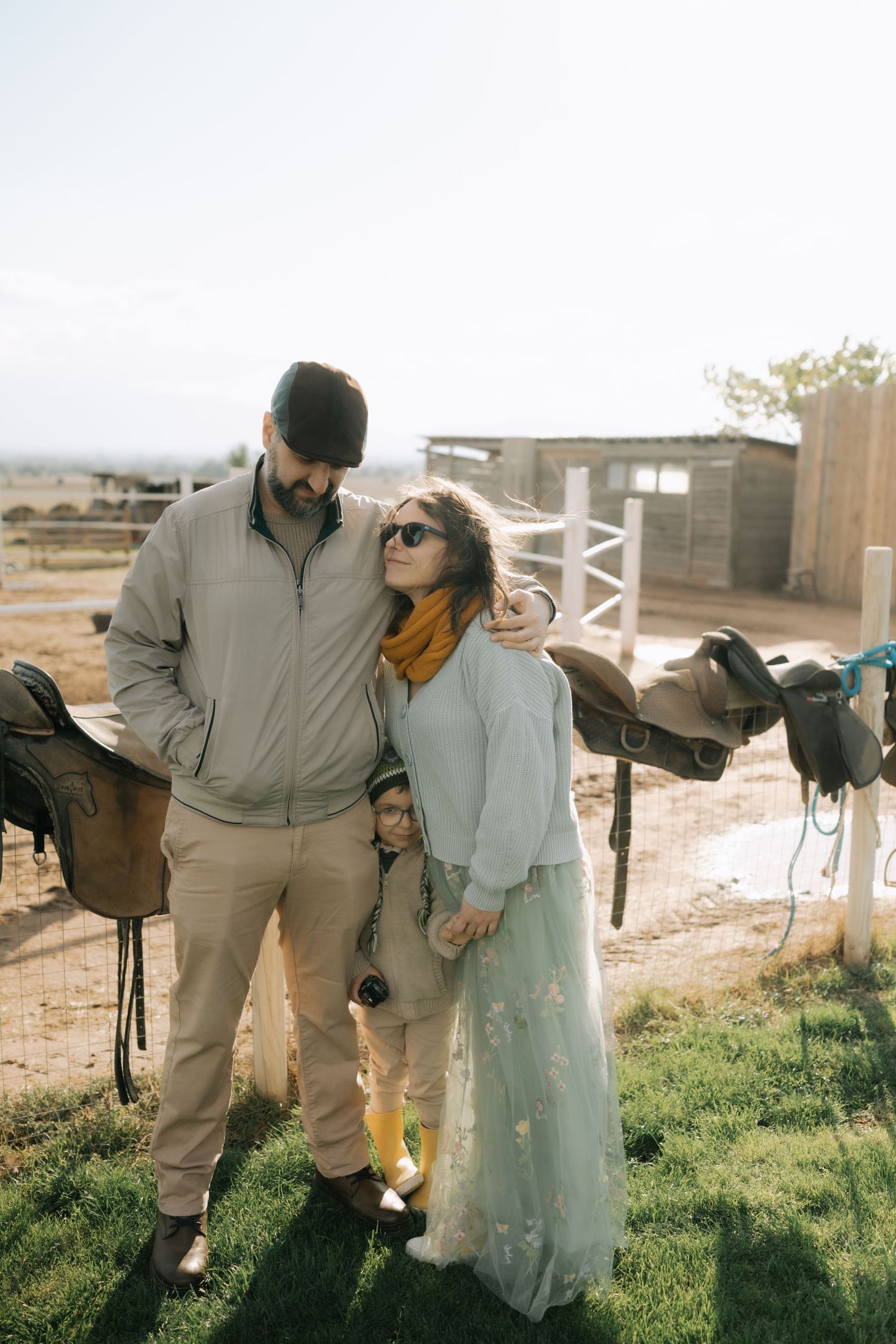 A couple stands together in casual attire on grassy farmland during golden hour lighting.