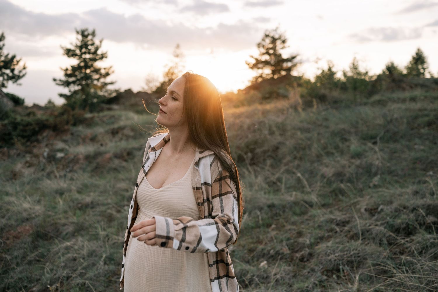 A person in a plaid shirt stands on a grassy hillside at sunset with evergreen trees in the background.