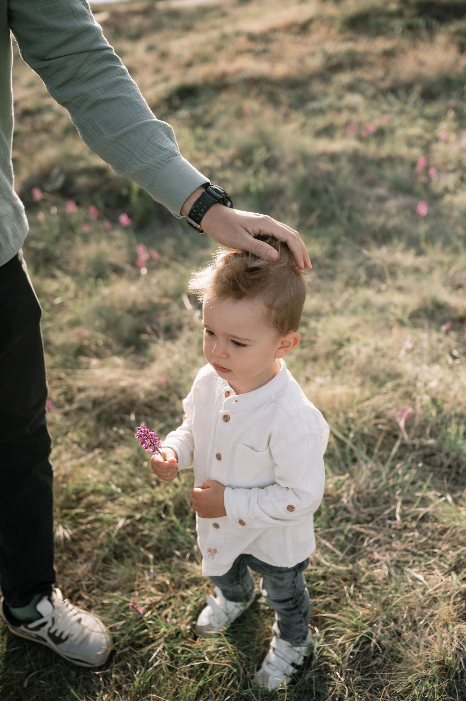 A toddler in a white shirt explores wildflowers in a grassy field while being guided by an adult's hand.