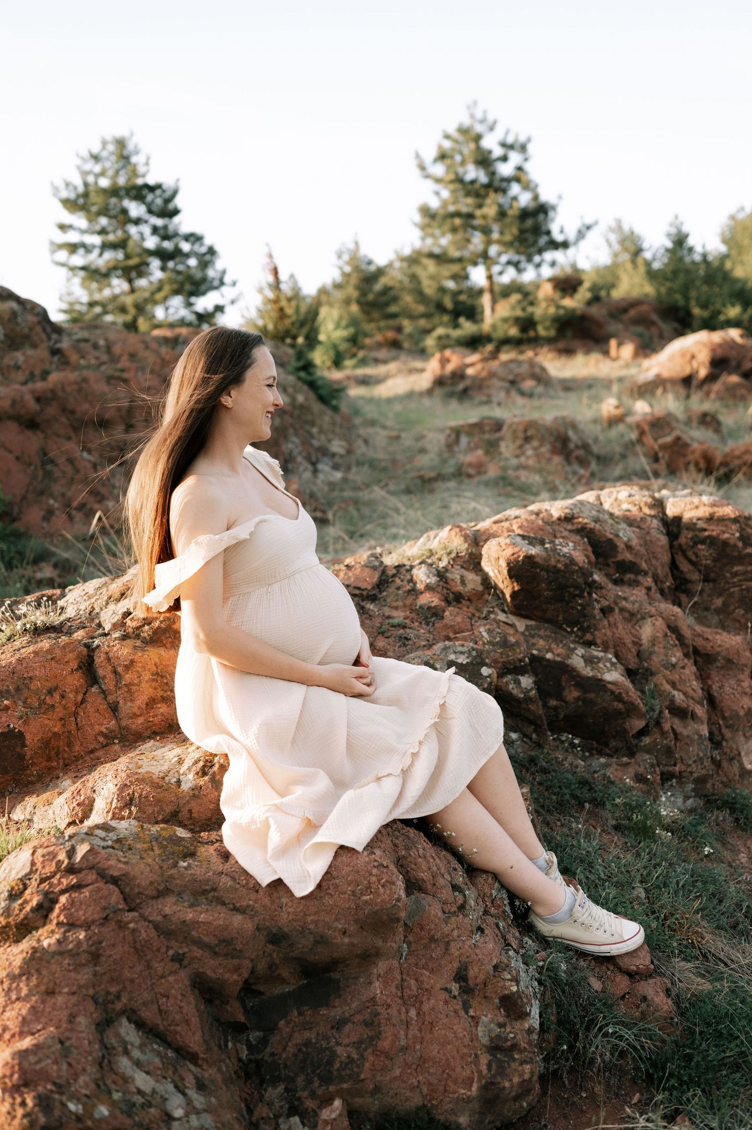 Person in a flowing white dress sits on rocky terrain during golden hour with evergreen trees in the background.