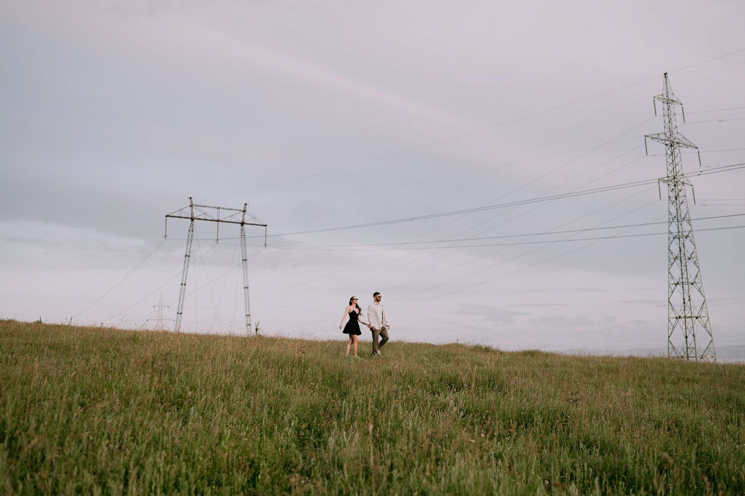 Two silhouetted figures walk along a grassy hilltop near power transmission lines under an overcast sky.