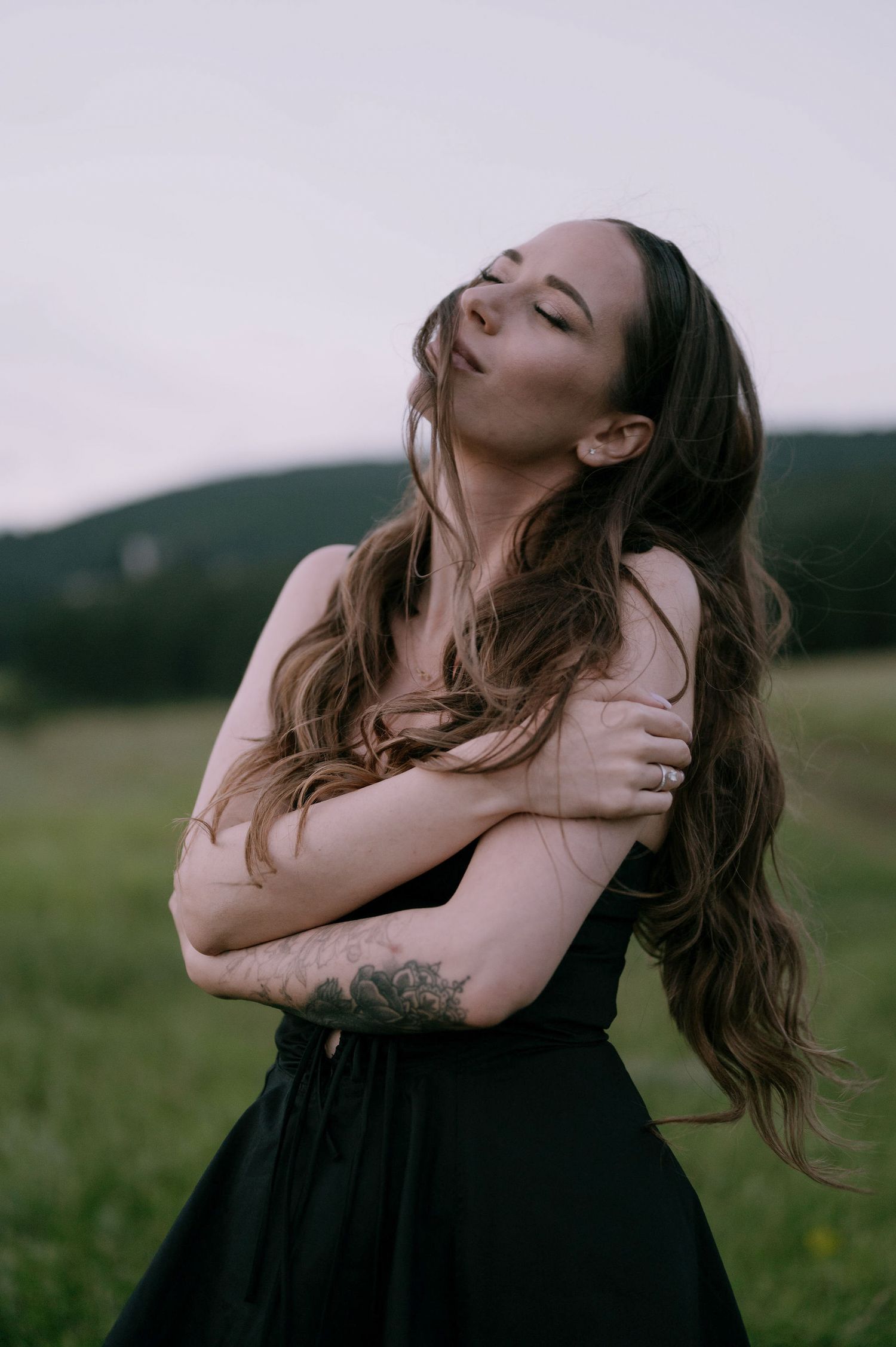 Woman in black dress poses expressively in a field with dramatic movements and flowing hair.